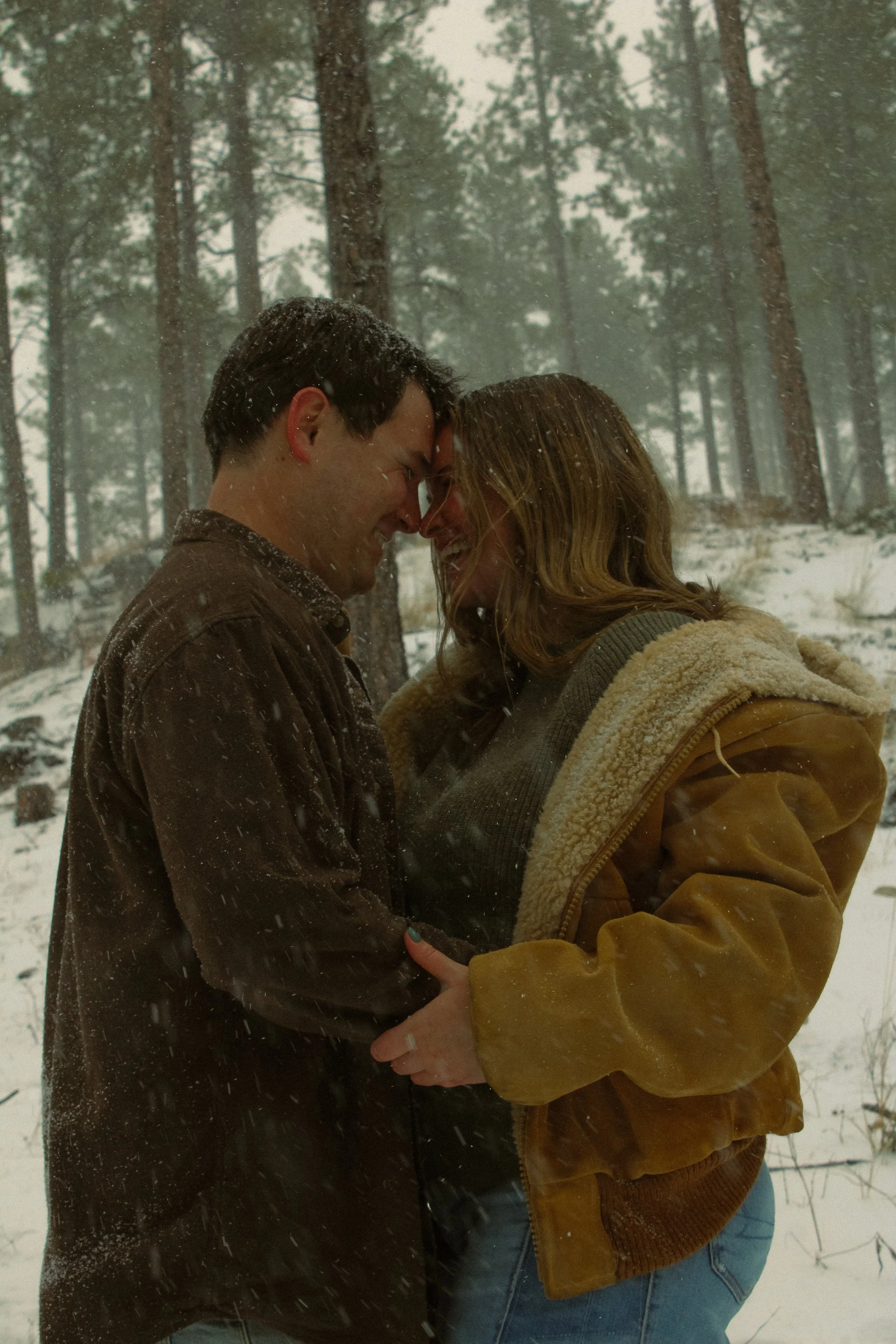 A couple is standing close together in a snowy forest with tall trees, their foreheads and noses touching, sharing an intimate moment while dressed in warm clothing.