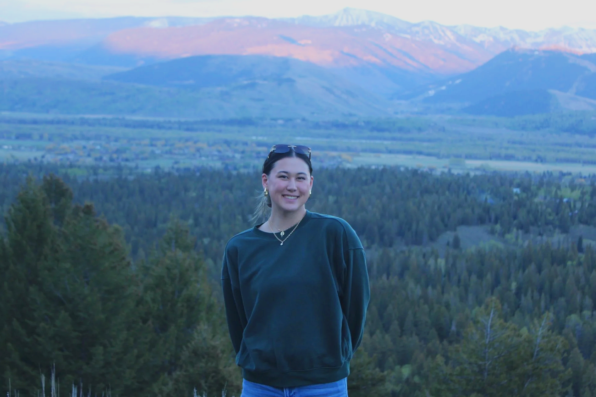 A young woman smiling outdoors with a mountain range in the background and trees in the foreground.