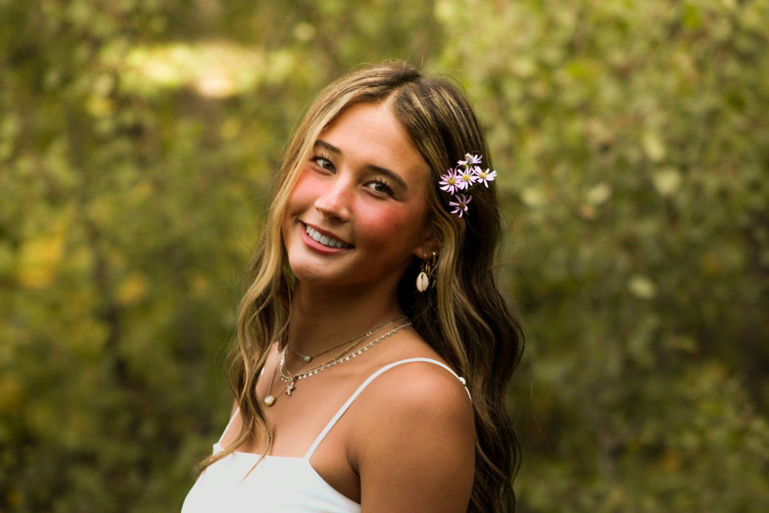 A woman with long wavy hair, smiling, wearing a white spaghetti strap top and multiple necklaces, with a small cluster of purple flowers tucked behind her ear, standing outdoors with a blurred green background of trees and foliage.