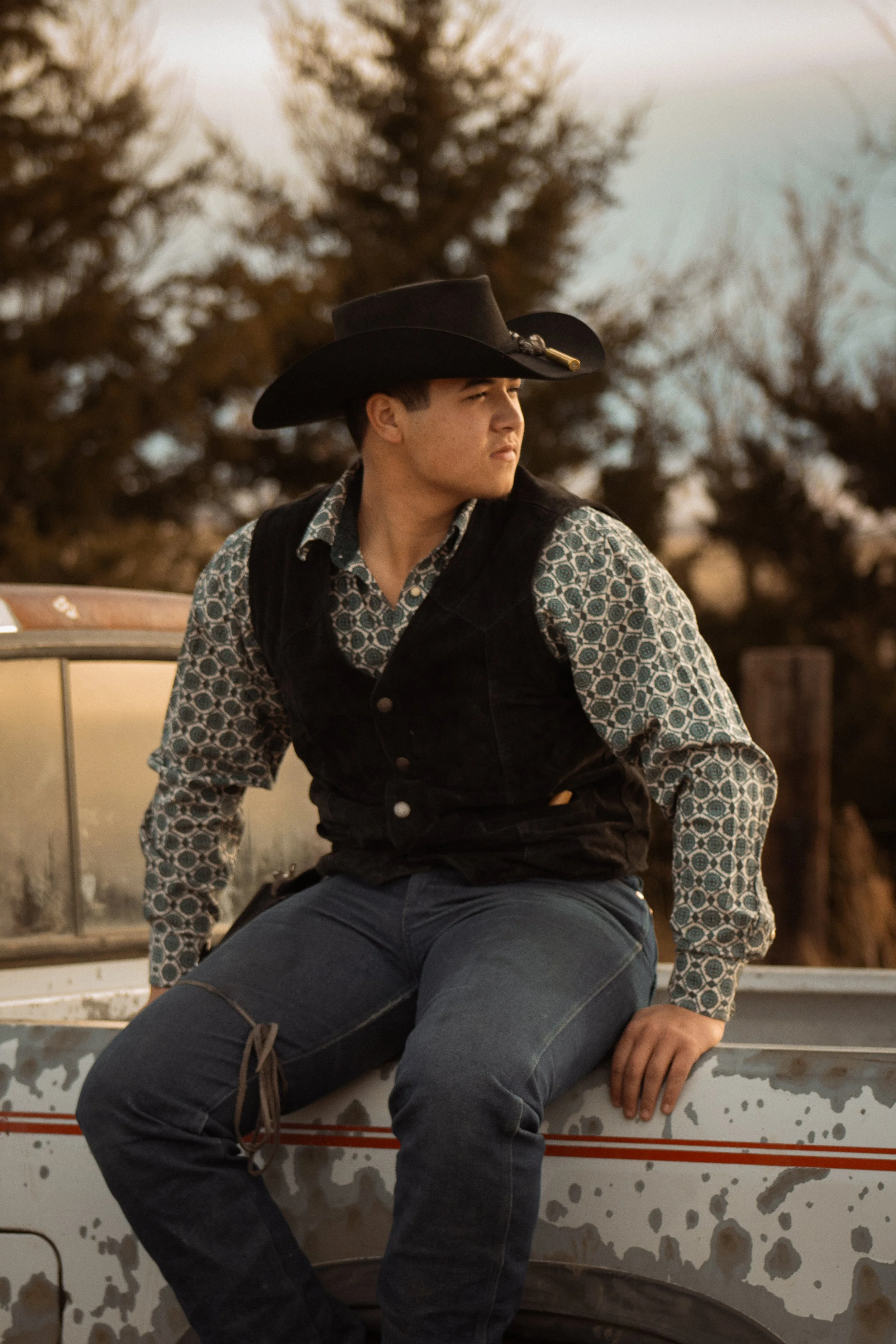 A young man sitting on a vintage truck wearing a black cowboy hat, patterned shirt, and vest, with trees in the background during sunset.