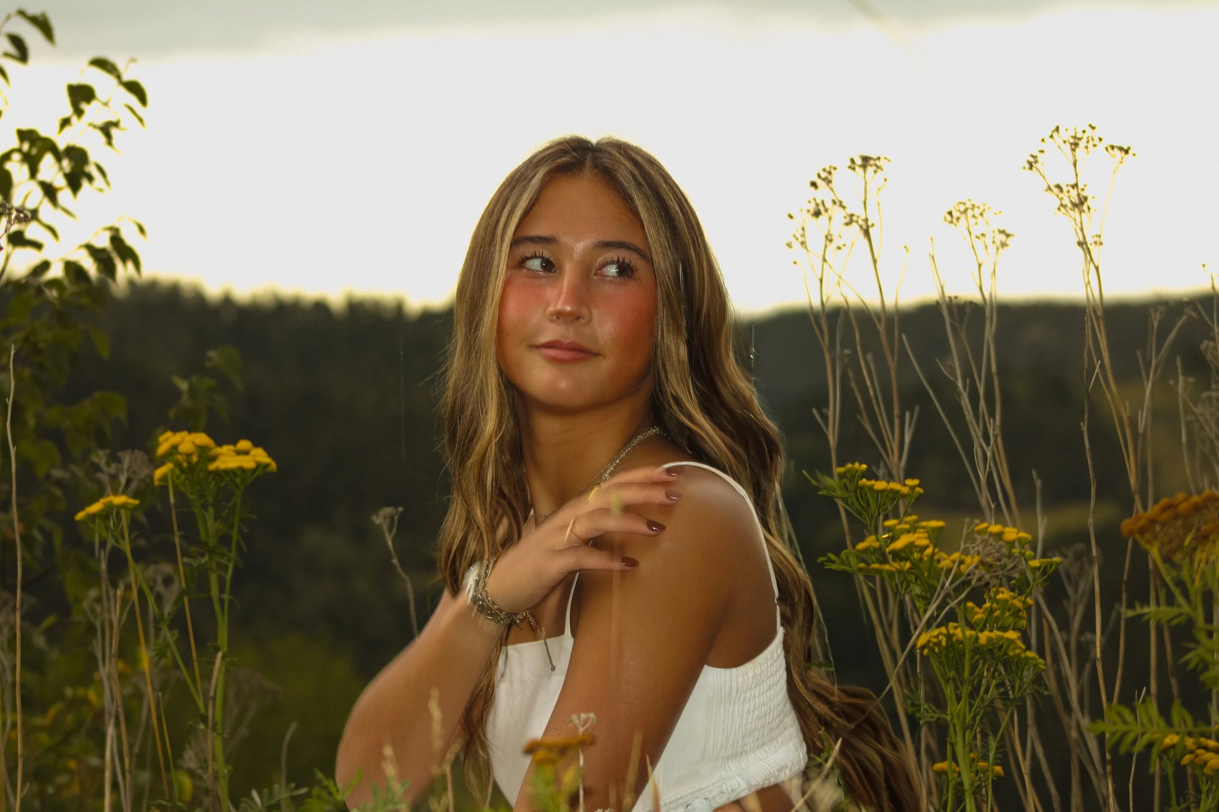 A young woman with long wavy hair standing in a field of yellow flowers during sunset, wearing a white sleeveless top and silver jewelry, looking to her left.