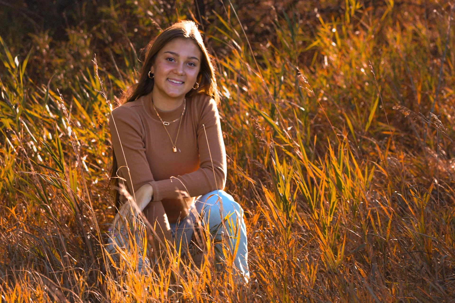 A young woman sitting outdoors among tall grass during sunset, smiling at the camera, wearing a brown long-sleeve shirt, gold jewelry, and light blue jeans.