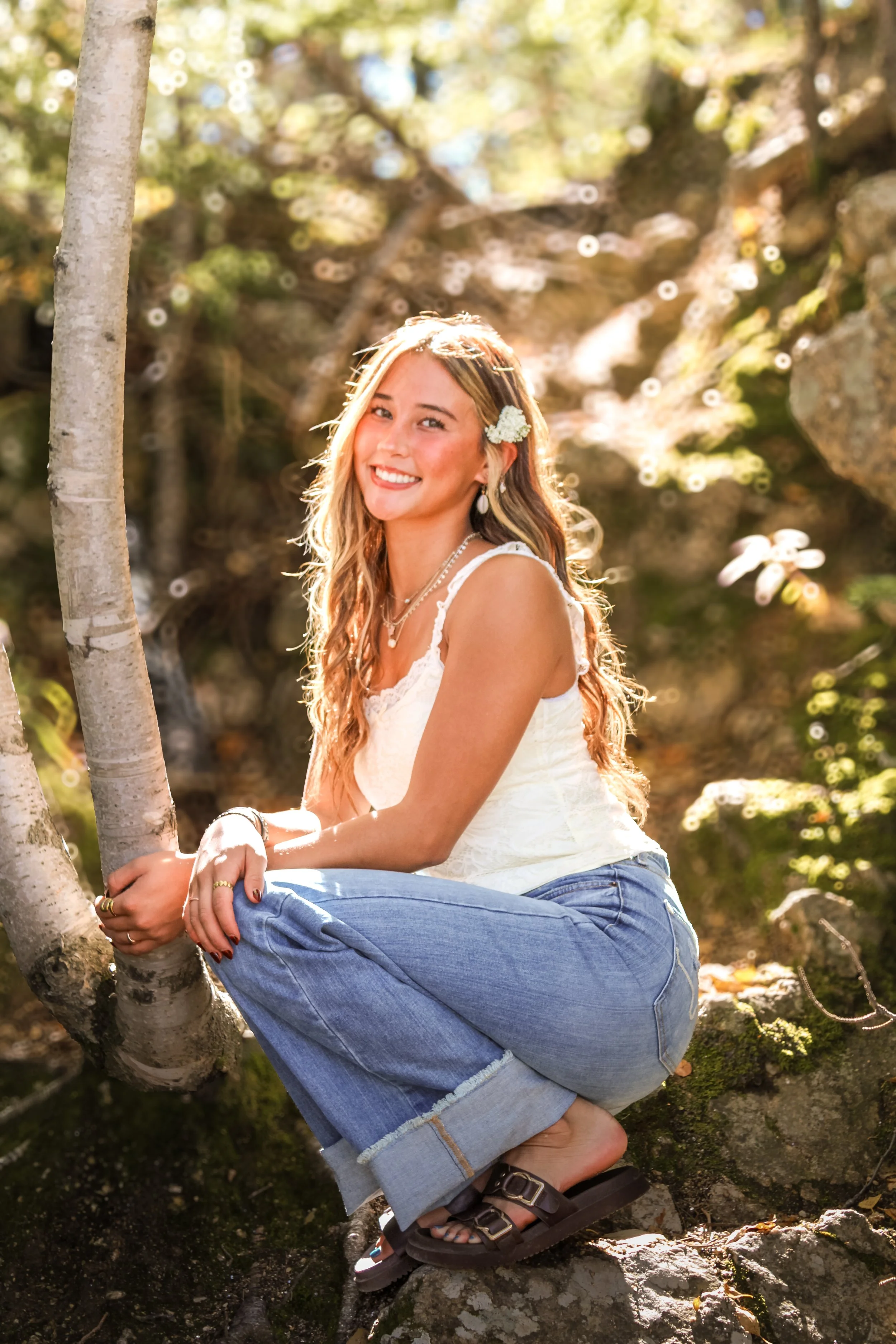 A woman with long blonde hair, wearing a white sleeveless top, blue jeans, and sandals, is crouching on a tree branch in a forest setting. She has a small white flower in her hair and is smiling at the camera, with sunlight filtering through the trees.