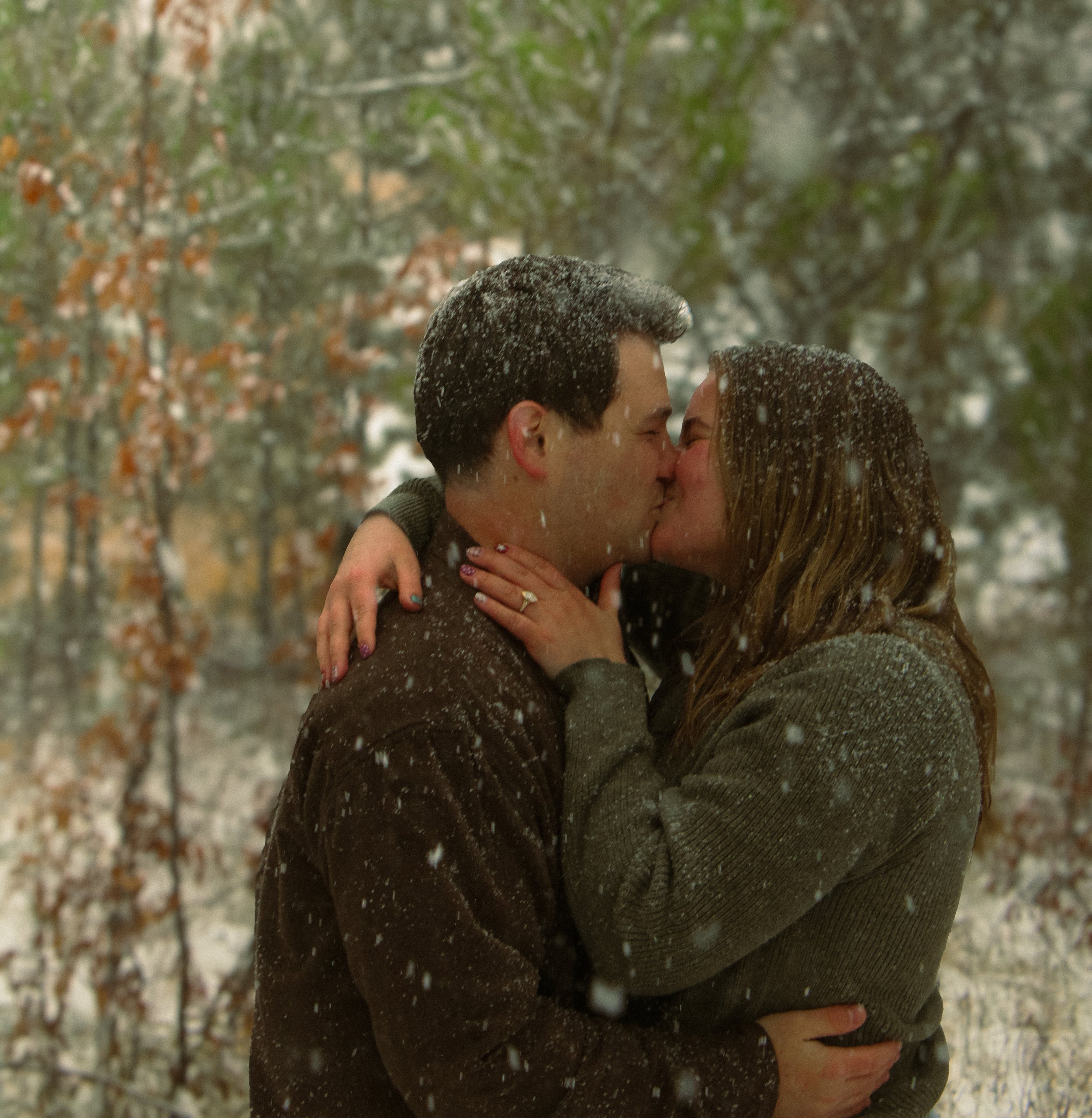 A close-up of a couple kissing outdoors in a snowy forest, with snowflakes on their hair and shoulders, surrounded by trees with snow-covered branches.