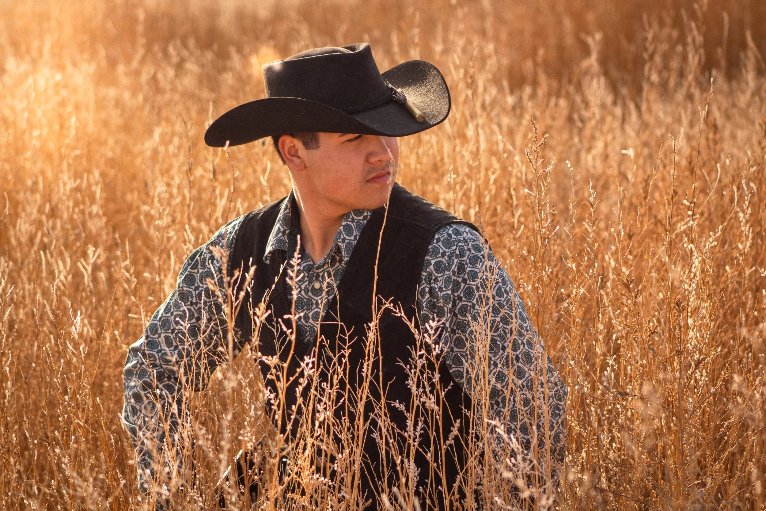 A young man wearing a black cowboy hat and patterned shirt standing in a golden field of tall dry grass, looking to the side.