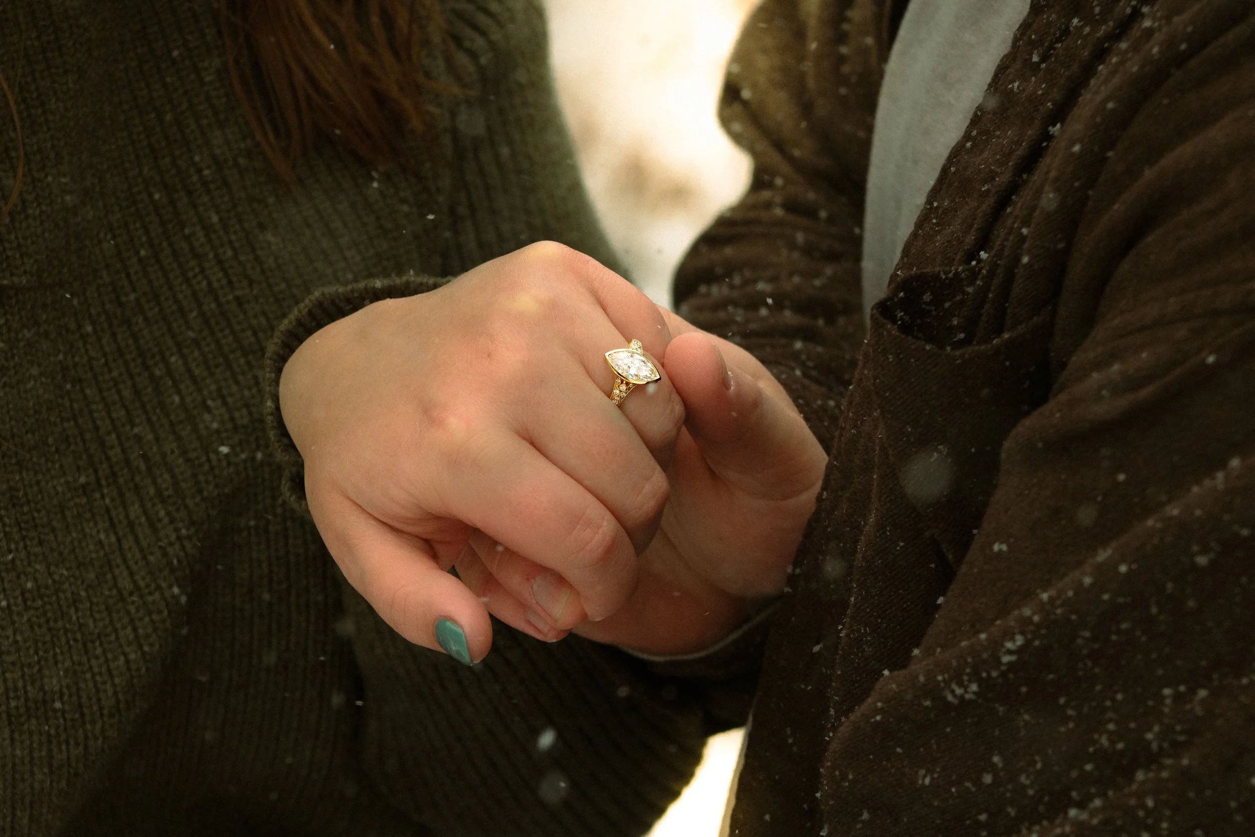 A person wearing a brown jacket and gray shirt is holding hands with another person. The person with the brown jacket is wearing a gold ring with a large, clear gemstone. The background is blurred.