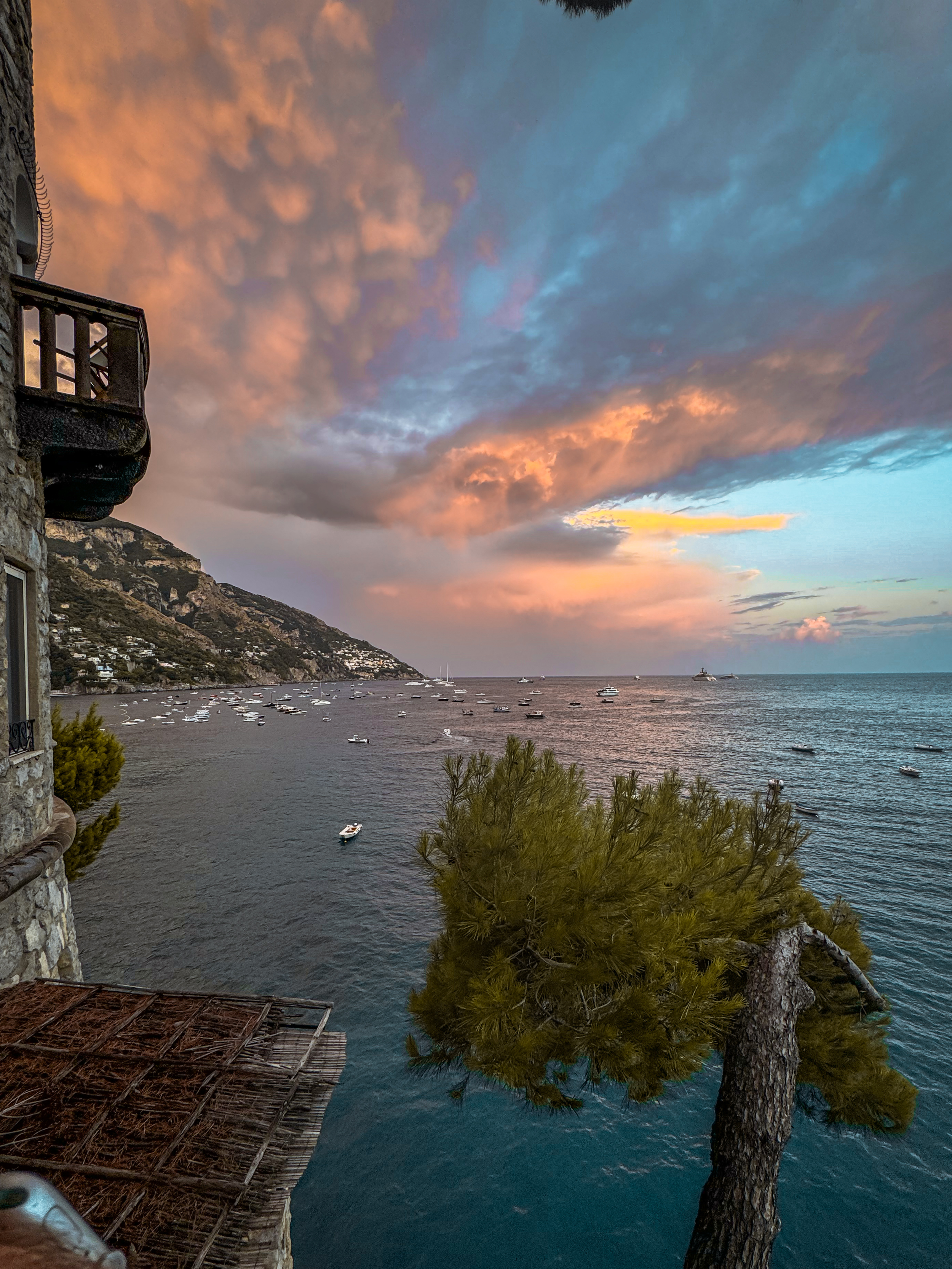 Sunset over a coastal village with boats anchored in the water, trees, and hillside houses in the distance.