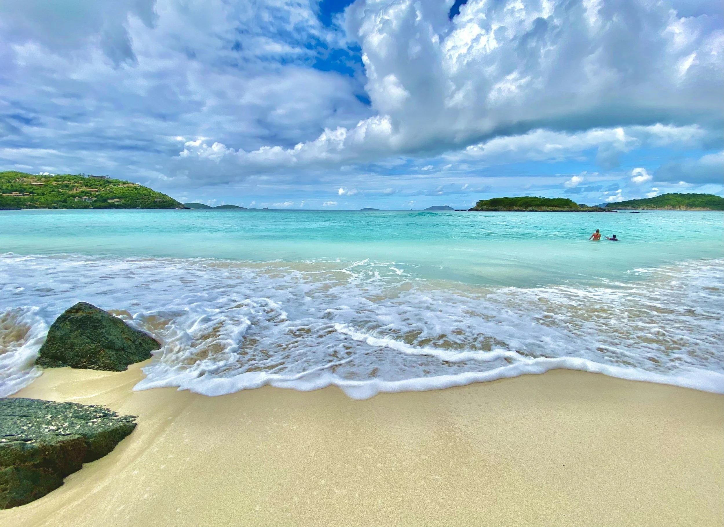 A beach scene with sand, rocks, and ocean waves. In the background, there are green hills and islands. The sky is partly cloudy with blue sky peeking through.