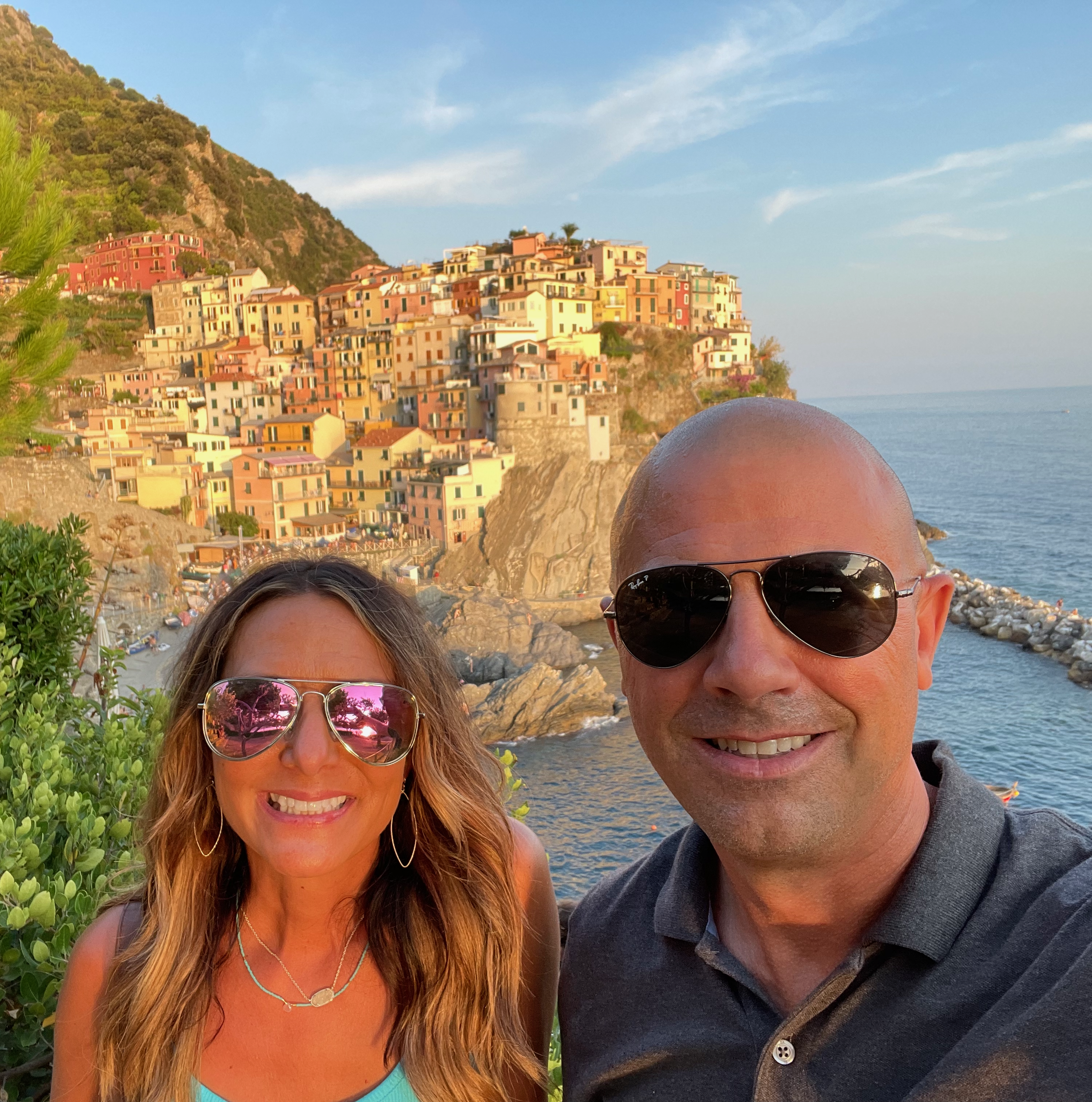 Close-up of a smiling woman with long hair wearing sunglasses, and a smiling man with sunglasses, behind a scenic coastal town with colorful buildings on a hillside, overlooking the ocean on a sunny day.
