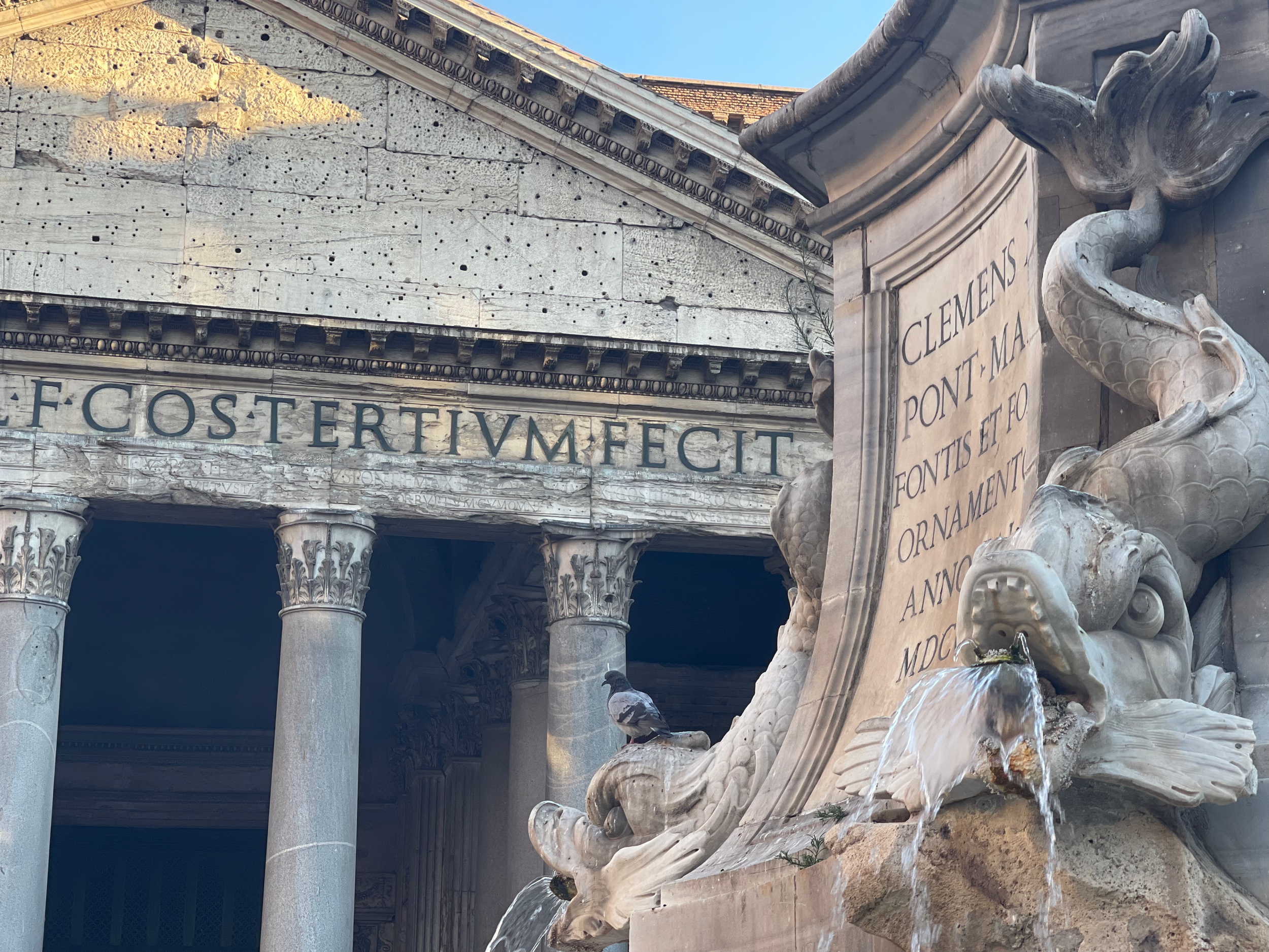 Close-up view of ancient Roman architecture, featuring weathered marble columns, a damaged pediment with Latin inscriptions, and ornamental sculptures including a sea creature. The sky is blue in the background.