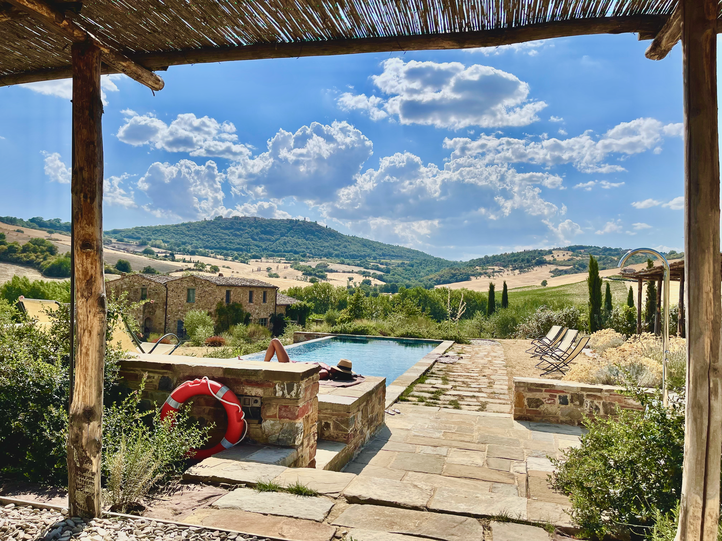 View of a scenic countryside landscape with rolling hills, a stone house, a swimming pool, and a clear sky with scattered clouds, seen from under a rustic wooden and bamboo pergola.