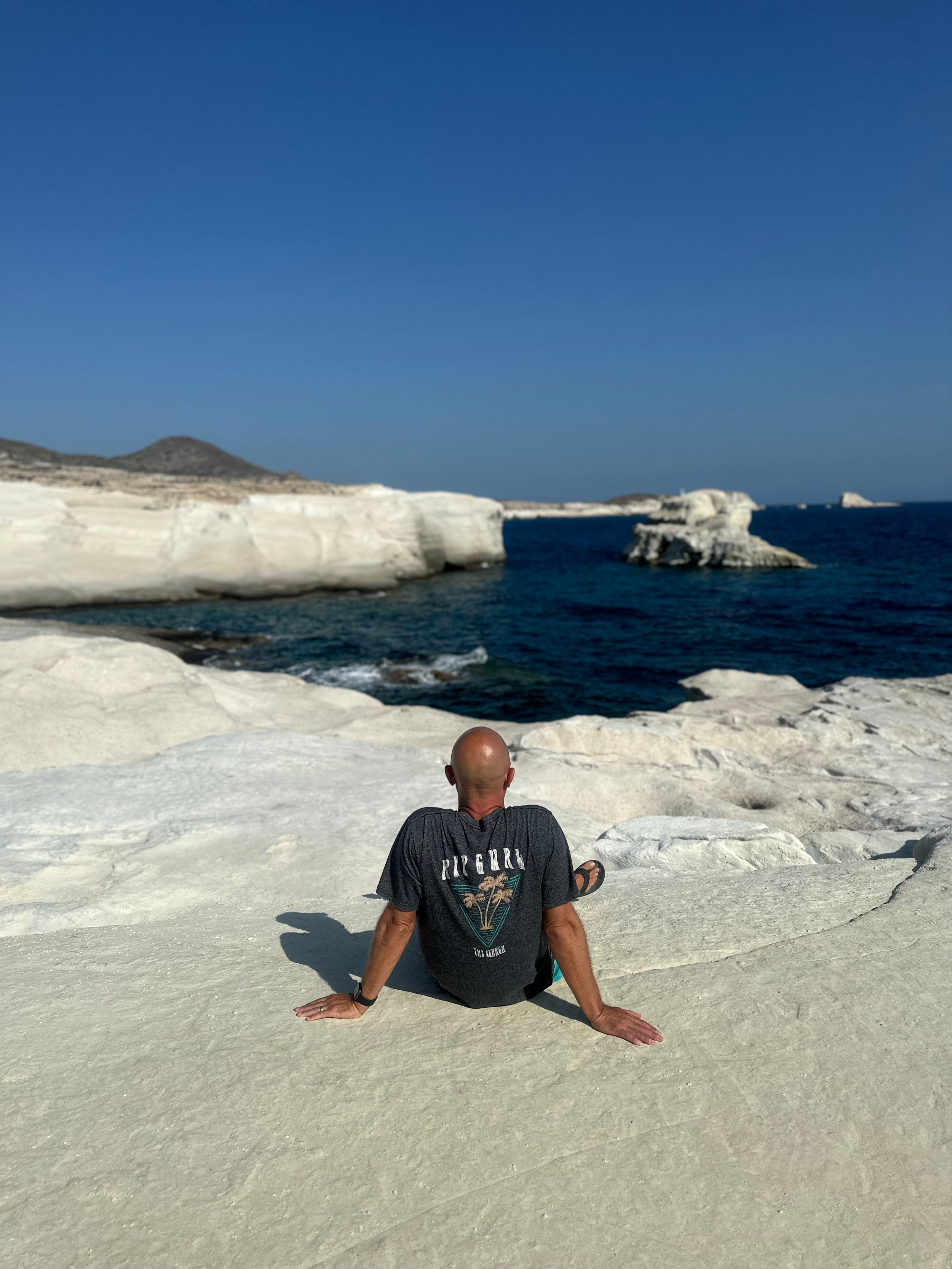 A man sitting on white rocky terrain near the sea, with white cliffs and a distant island under a clear blue sky.