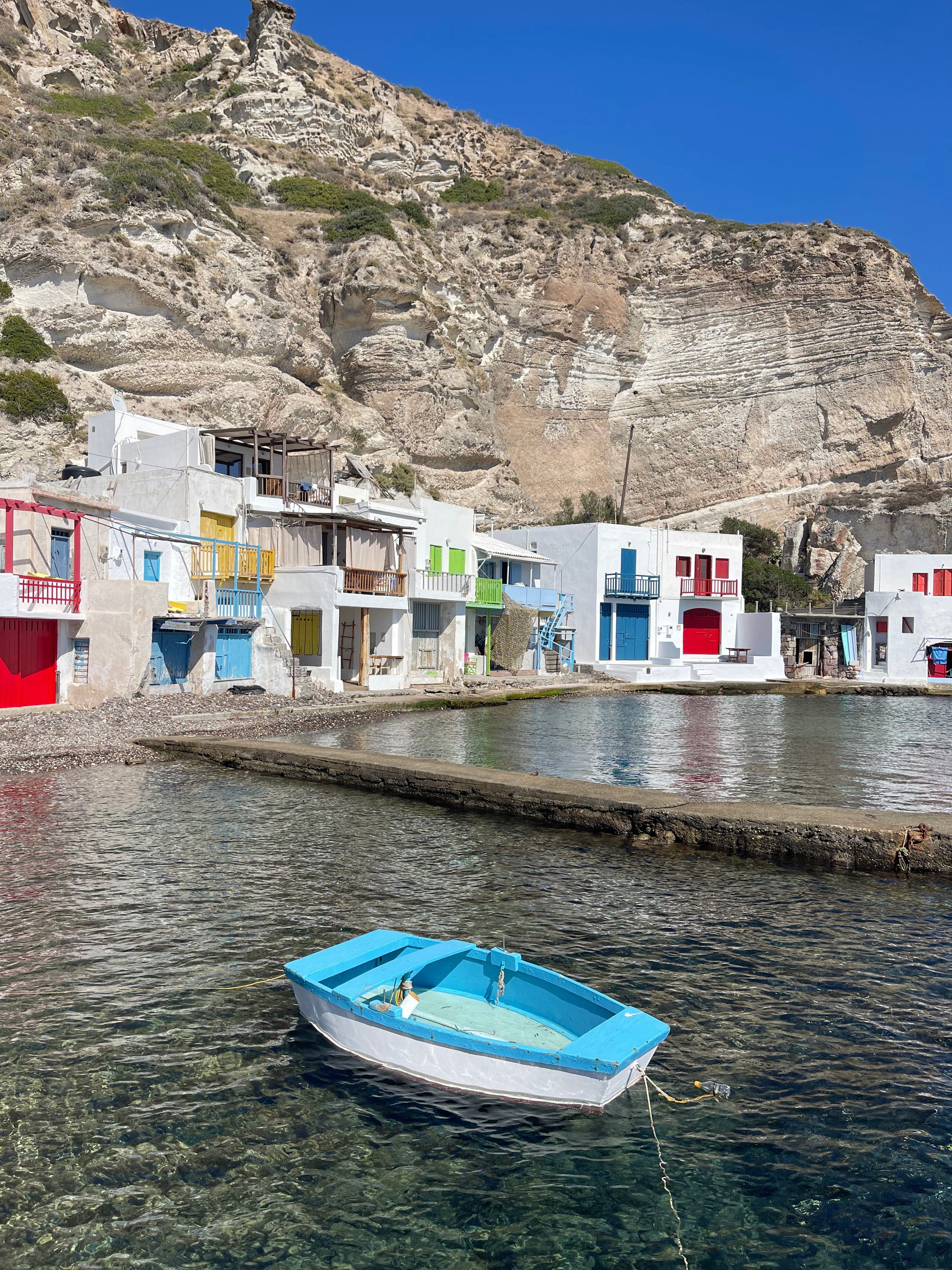 A small blue boat floating on clear water with pebbles visible beneath, in front of a row of white buildings with colorful doors and balconies at the water's edge, and a large rocky mountain behind them under a bright blue sky.