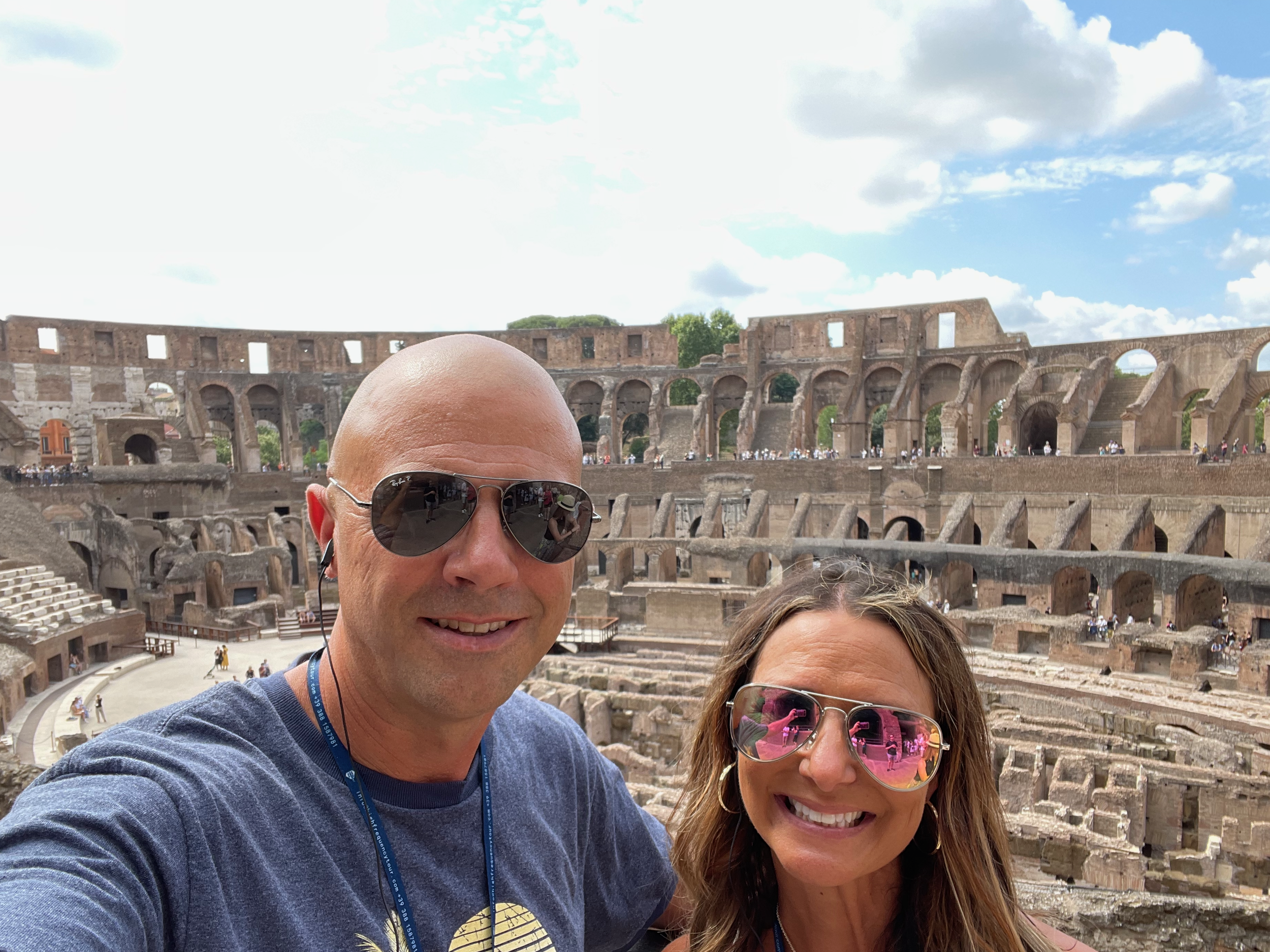 Close-up of a man and woman taking a selfie at the Roman Colosseum with the ancient amphitheater in the background, under a partly cloudy sky.