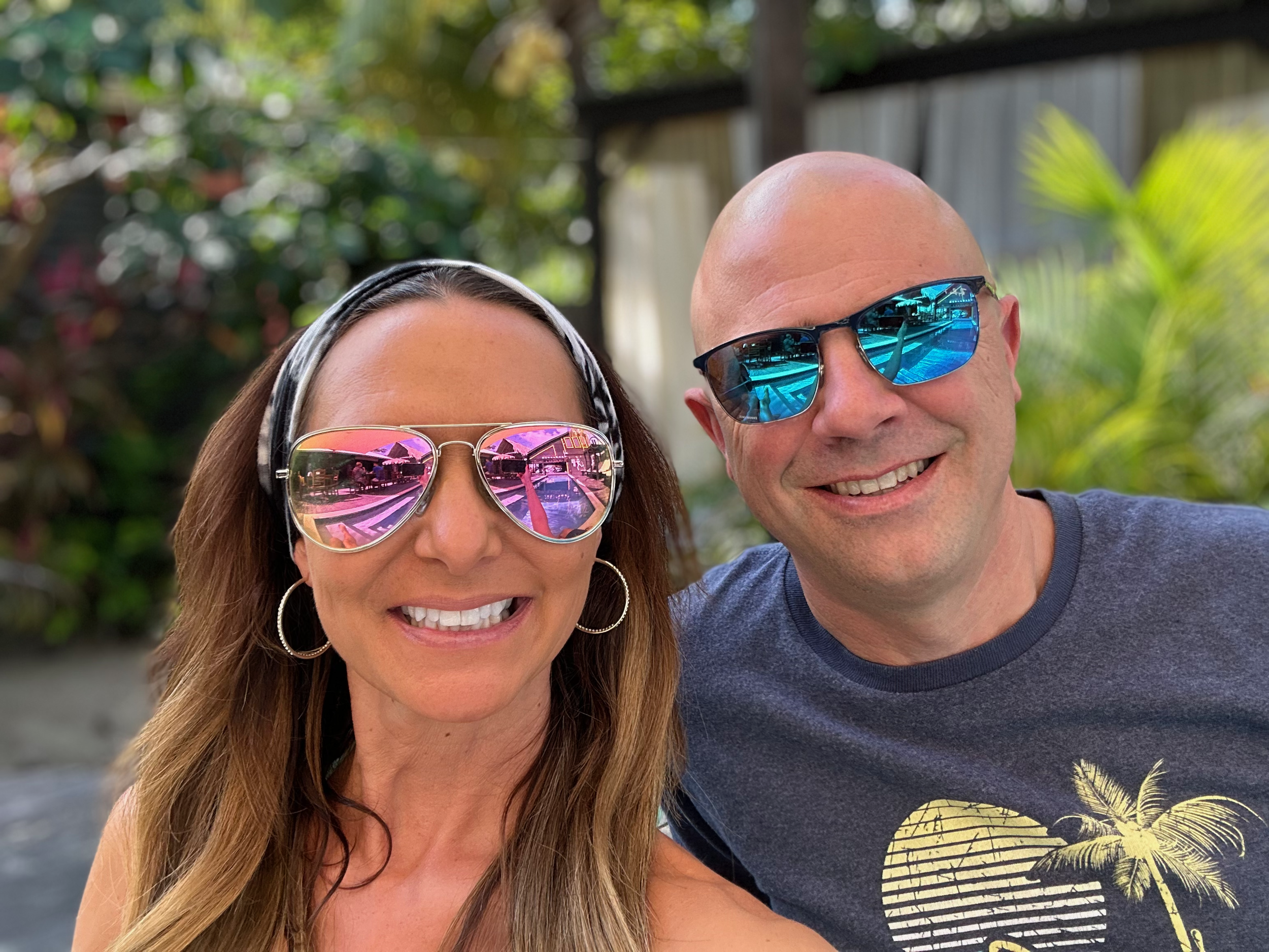 A woman with long hair and a striped headband wearing reflective pink sunglasses, smiling, next to a bald man wearing blue sunglasses, smiling, outdoors with green foliage in the background.