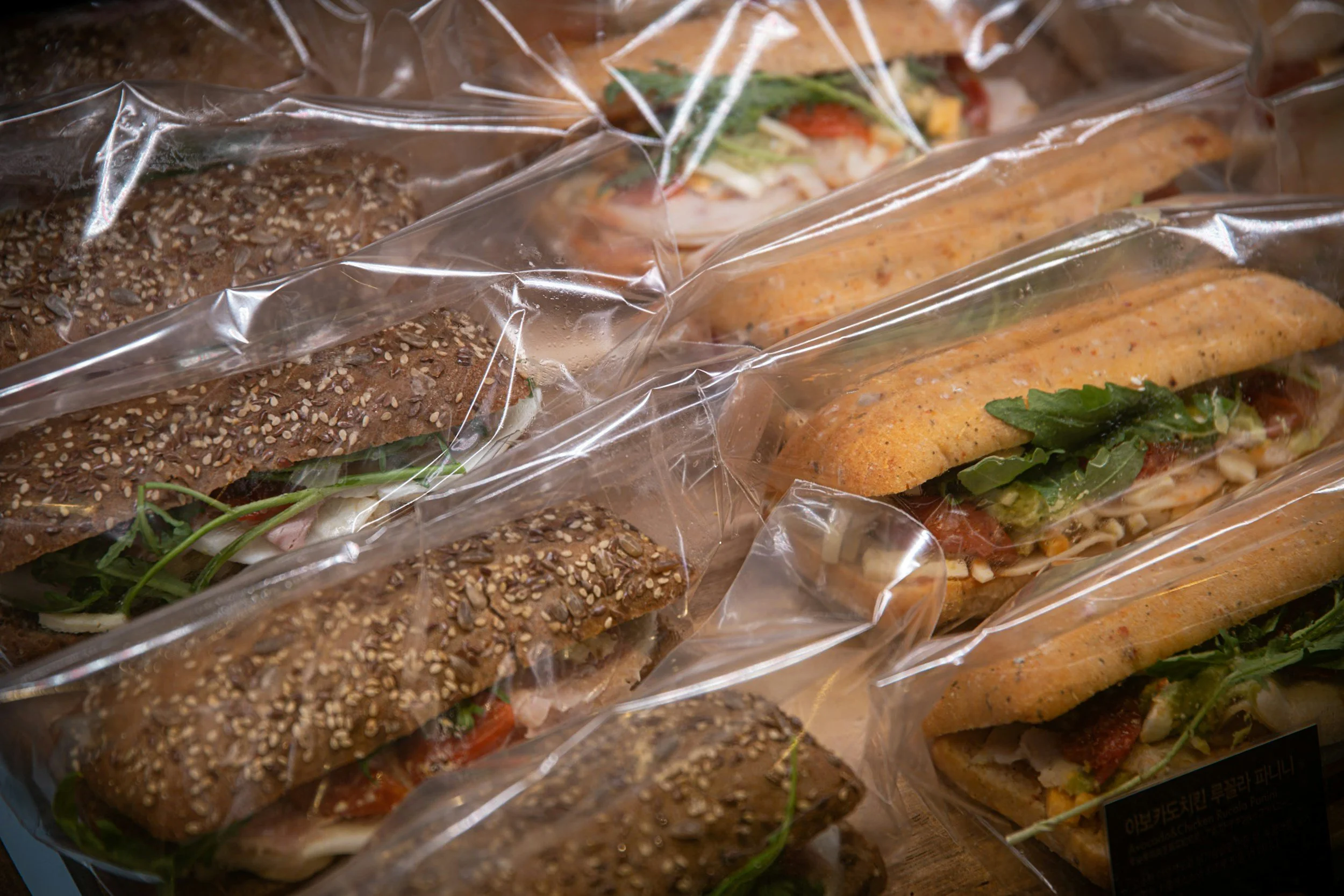 assorted sandwiches wrapped in clear plastic, featuring various types of bread such as whole grain with seeds and baguettes, with visible fillings like greens and meats.