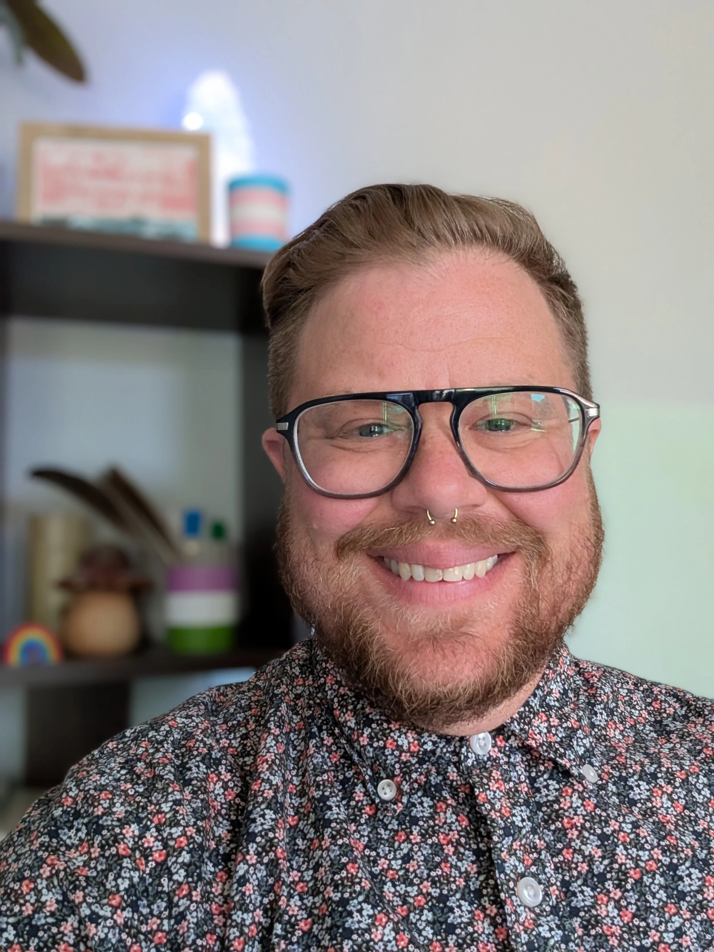 A smiling person with short brown hair, glasses, a septum piercing, and a beard wearing a floral patterned shirt in an indoor setting.