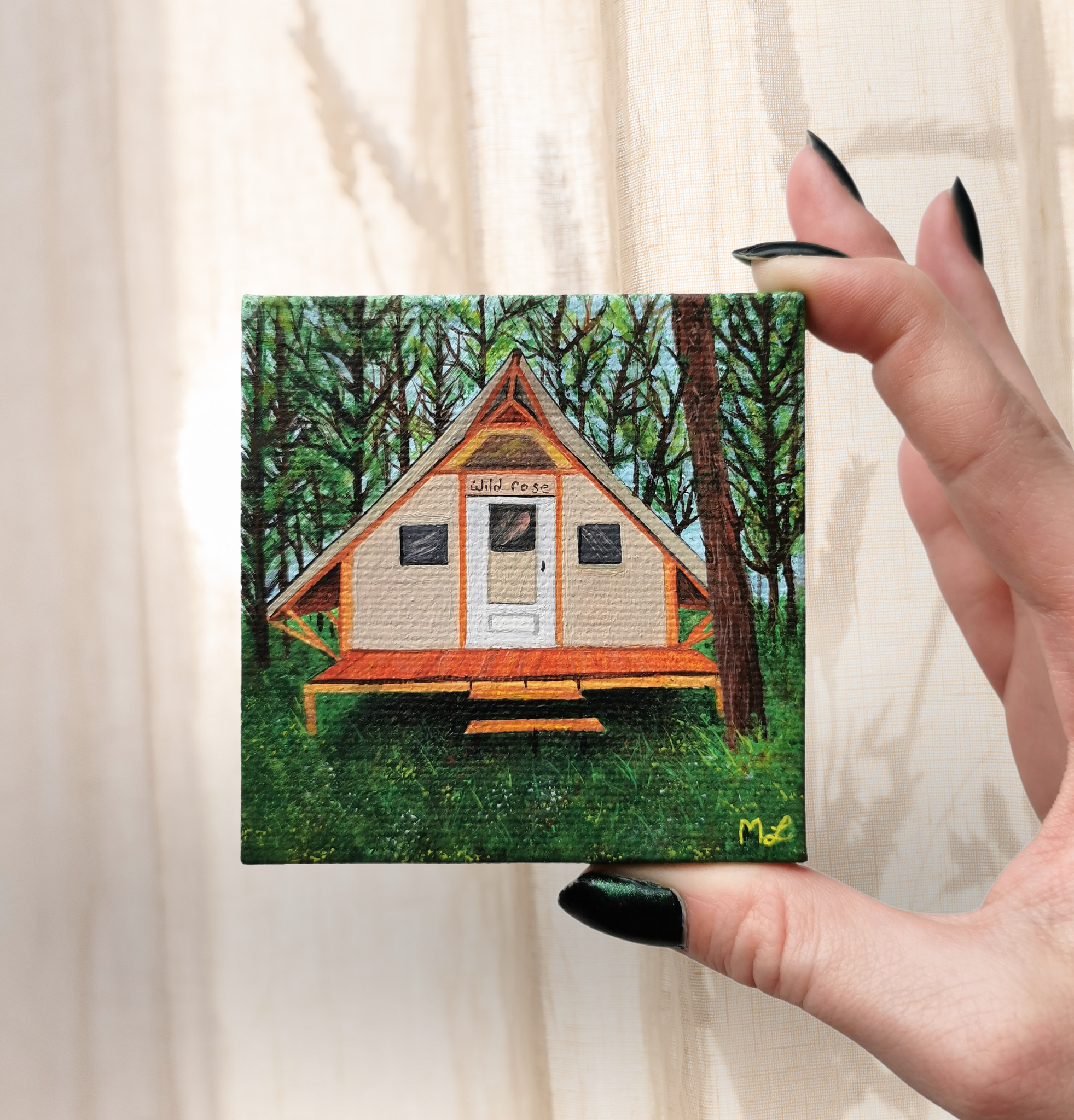 Small painting of a tiny house with a white door and dark windows surrounded by trees, with the words 'tiny house' written above the door, being held by a person with black painted nails near a light curtain background.