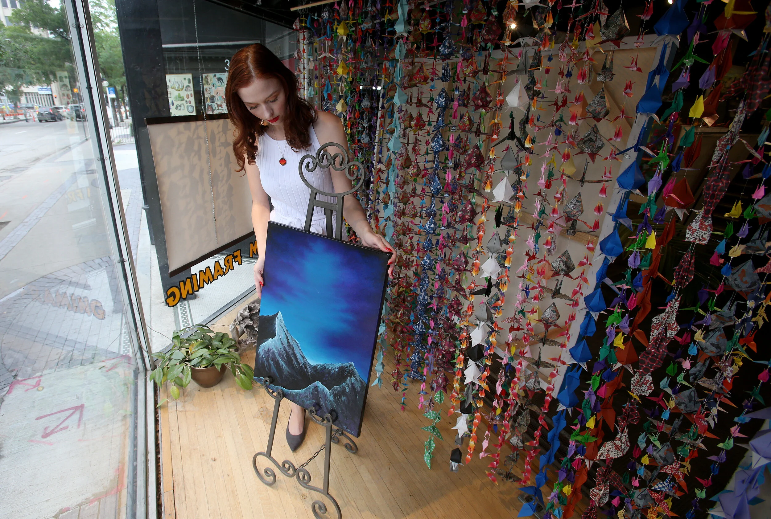 A woman (the artist, Margie Lucier) with long red hair and a white dress looking at a mountain landscape painting inside an art store, surrounded by colorful origami decorations.