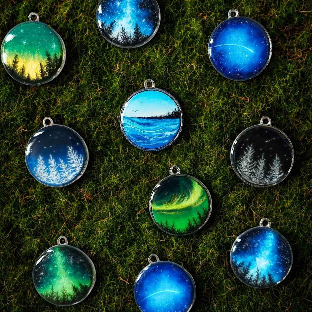 Collection of round pendants with nature and night sky paintings, arranged on a mossy surface.