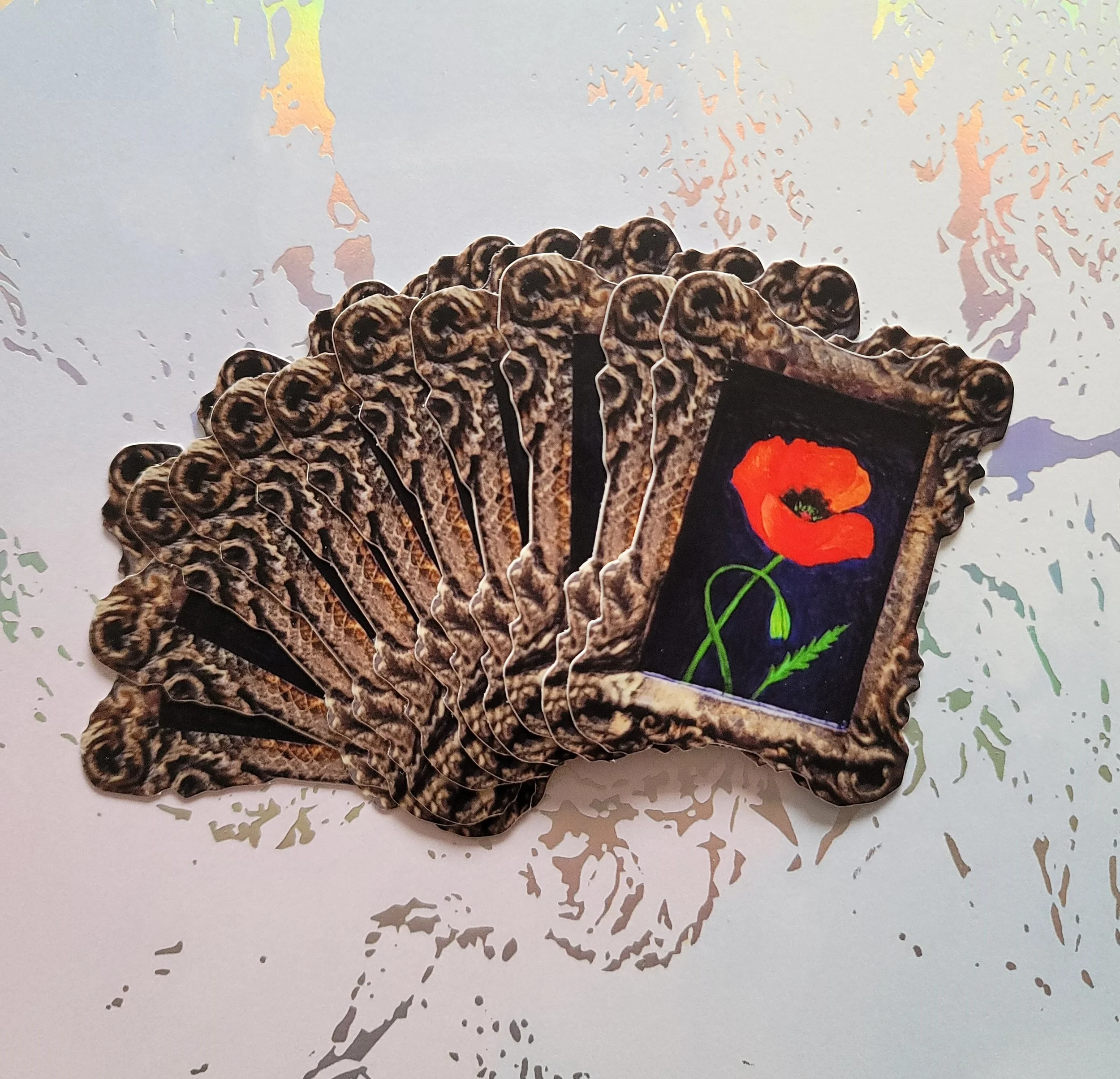 A stack of stickers, with a bright red poppy flower on it, arranged on a white surface with a splattered paint pattern.