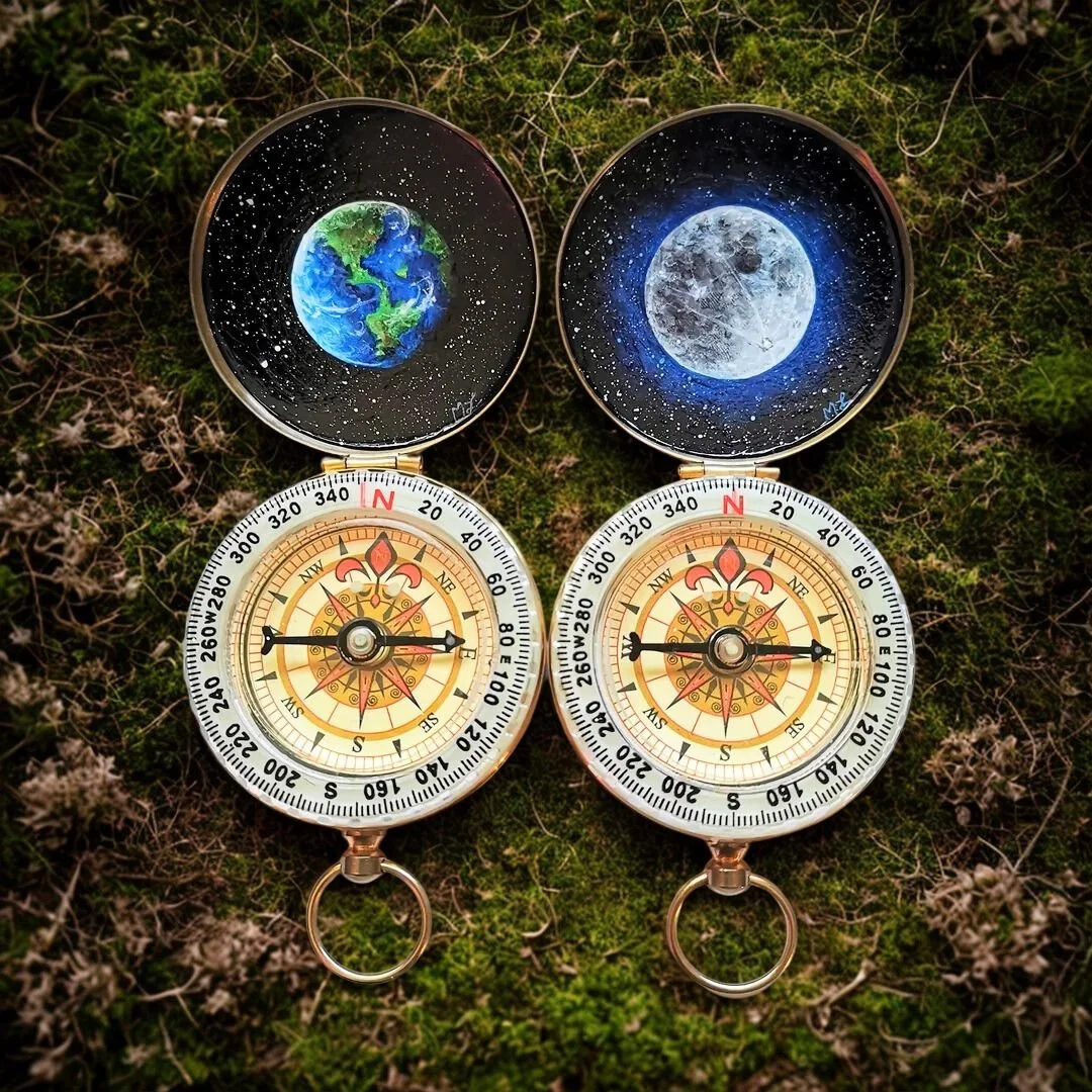 Two compasses with celestial artwork on lids, one depicting Earth and the other the Moon, placed on mossy ground.