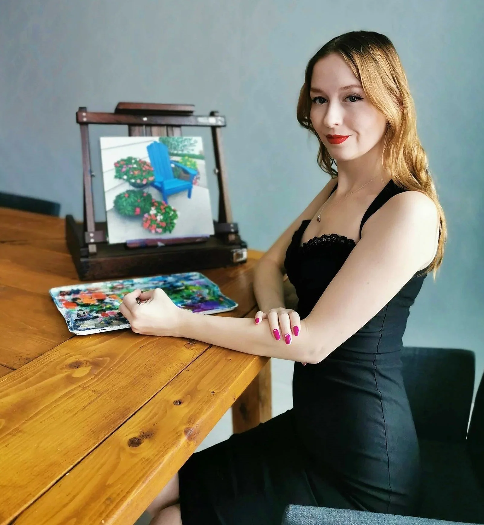 A woman with red hair and red lipstick is sitting at a wooden table (the artist Margie Lucier), with a painting with a colorful palette. There is an original painting of an outdoor scene with a blue chair and potted plants on the table.