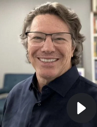 A man with glasses smiling in an indoor setting with bookshelves in the background.