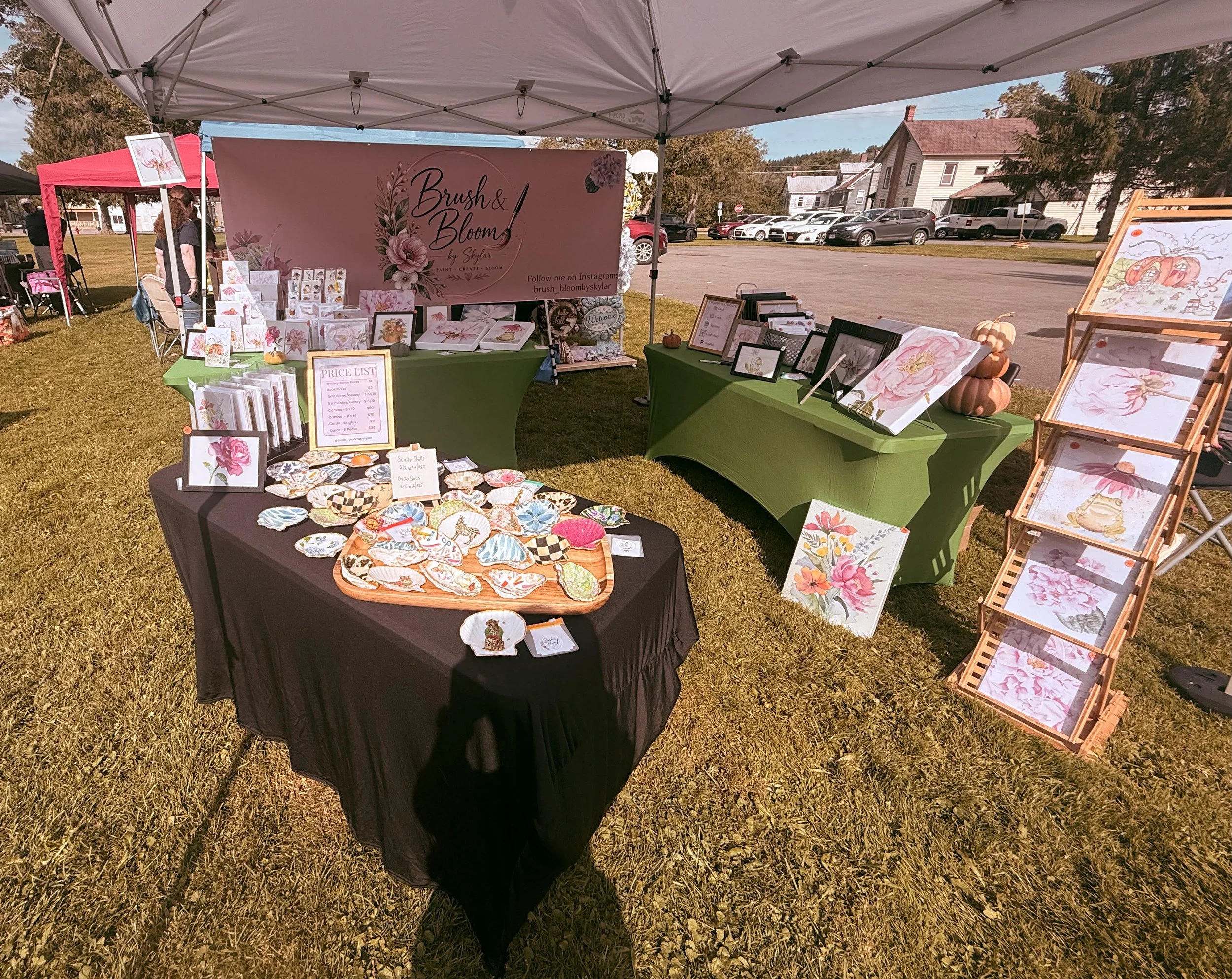 Outdoor craft fair booth with floral artwork, greeting cards, and decorative items displayed on tables and easels under white canopy tents, with a pink promotional banner that reads 'Brush & Bloom by Skylar'.