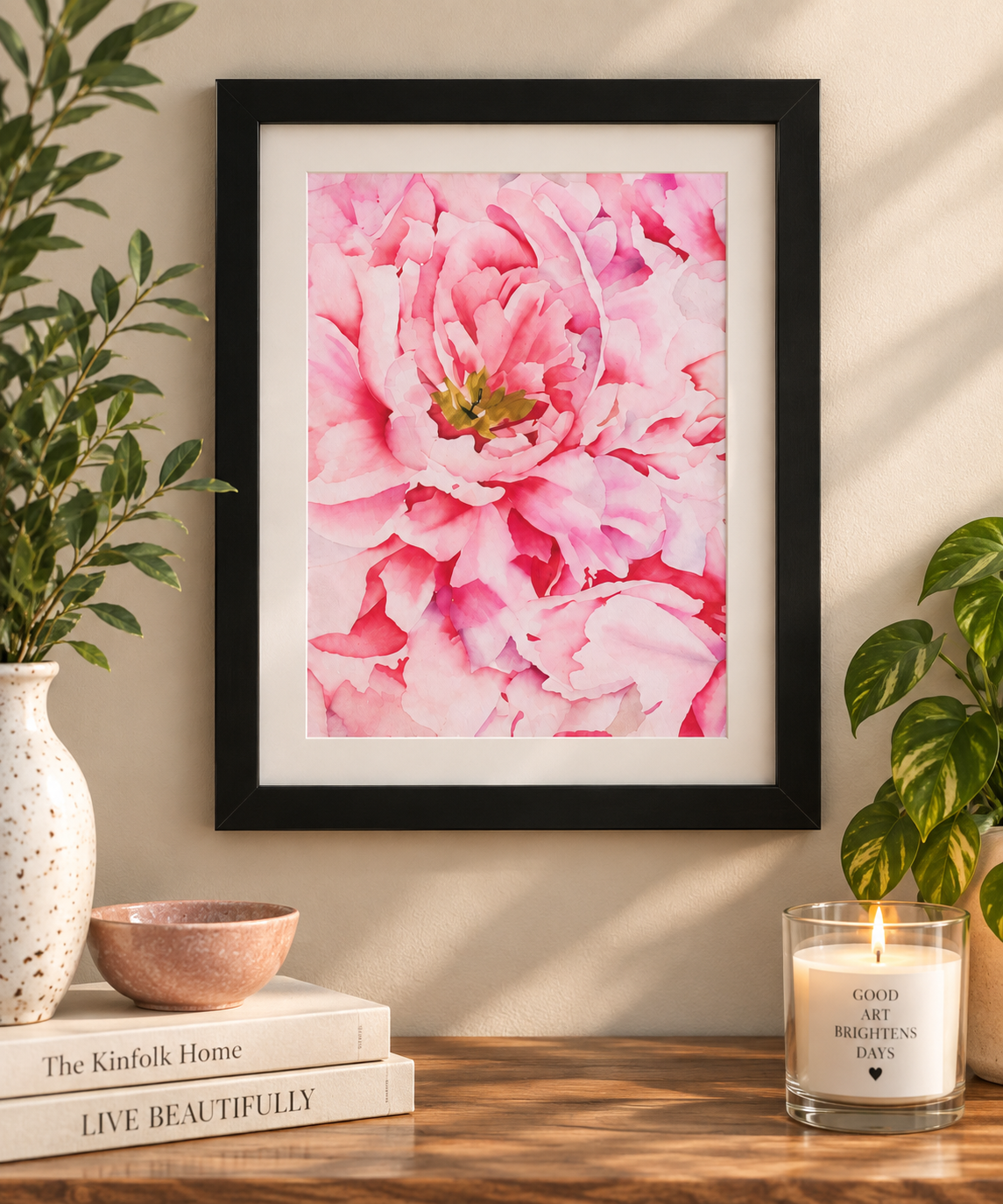 Pink peony watercolor print in a black frame with a white mat, displayed on a warm neutral wall with plants and cozy home décor.