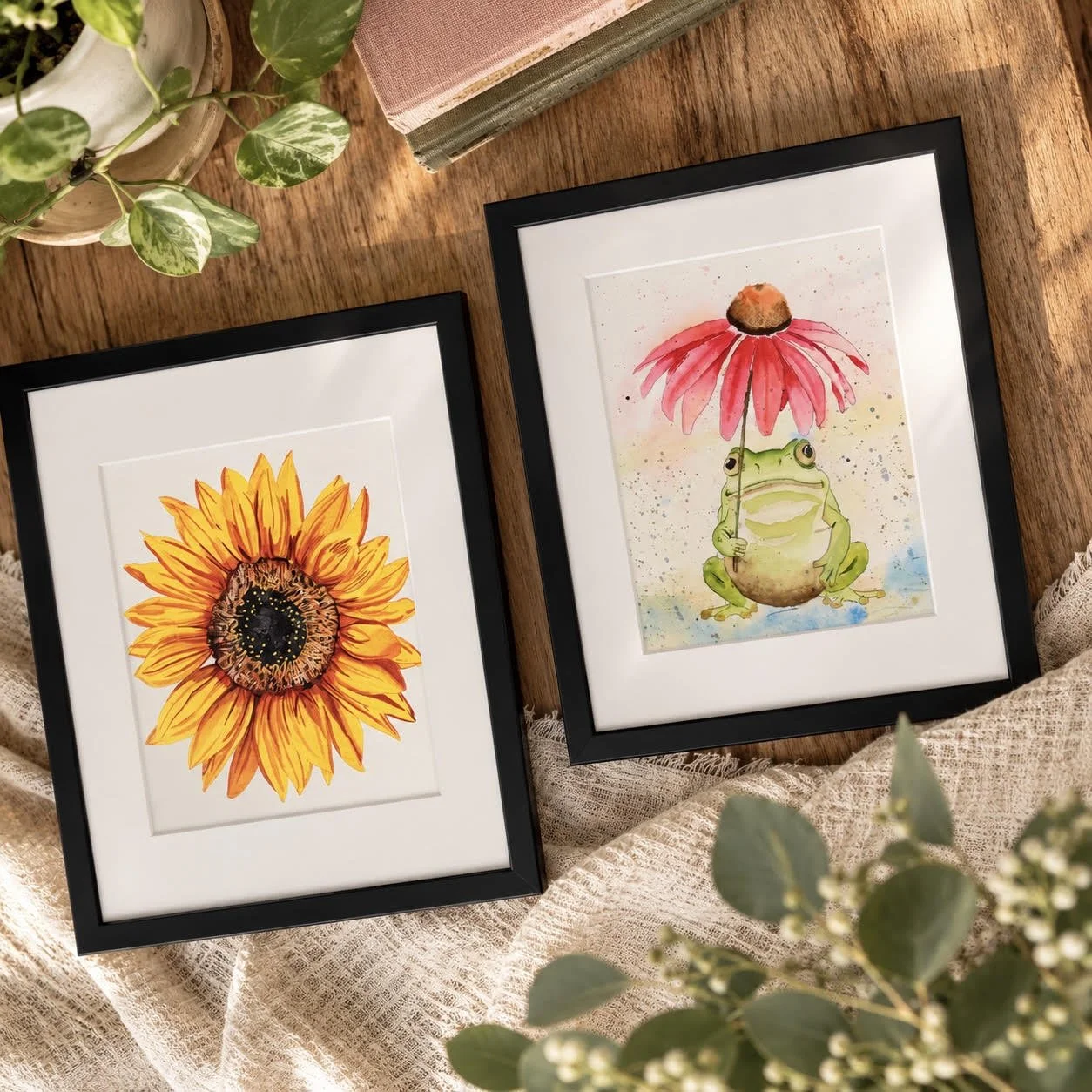 Two black-framed watercolor artworks displayed on a warm wooden surface, featuring a bright sunflower painting and a whimsical green frog beside a pink coneflower, styled with soft fabric, greenery, and neutral decor.