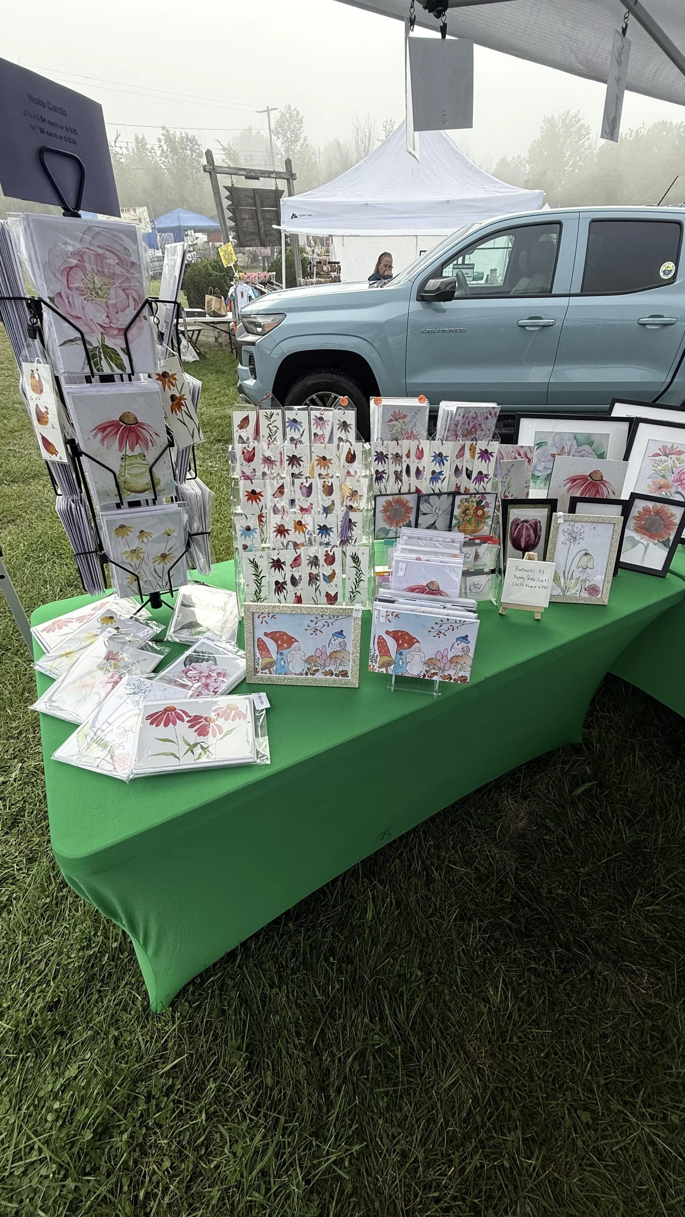 Outdoor craft fair booth with various floral-themed greeting cards and artwork displayed on a green table, with a blue truck and tents in the background.