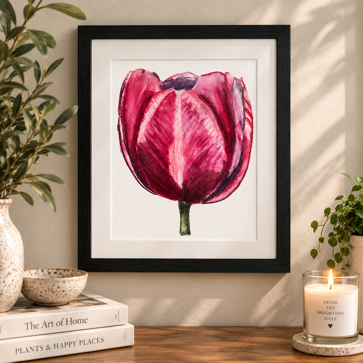 Framed watercolor print of a deep pink and burgundy tulip displayed in a black frame against a soft green wall with botanical studio decor.