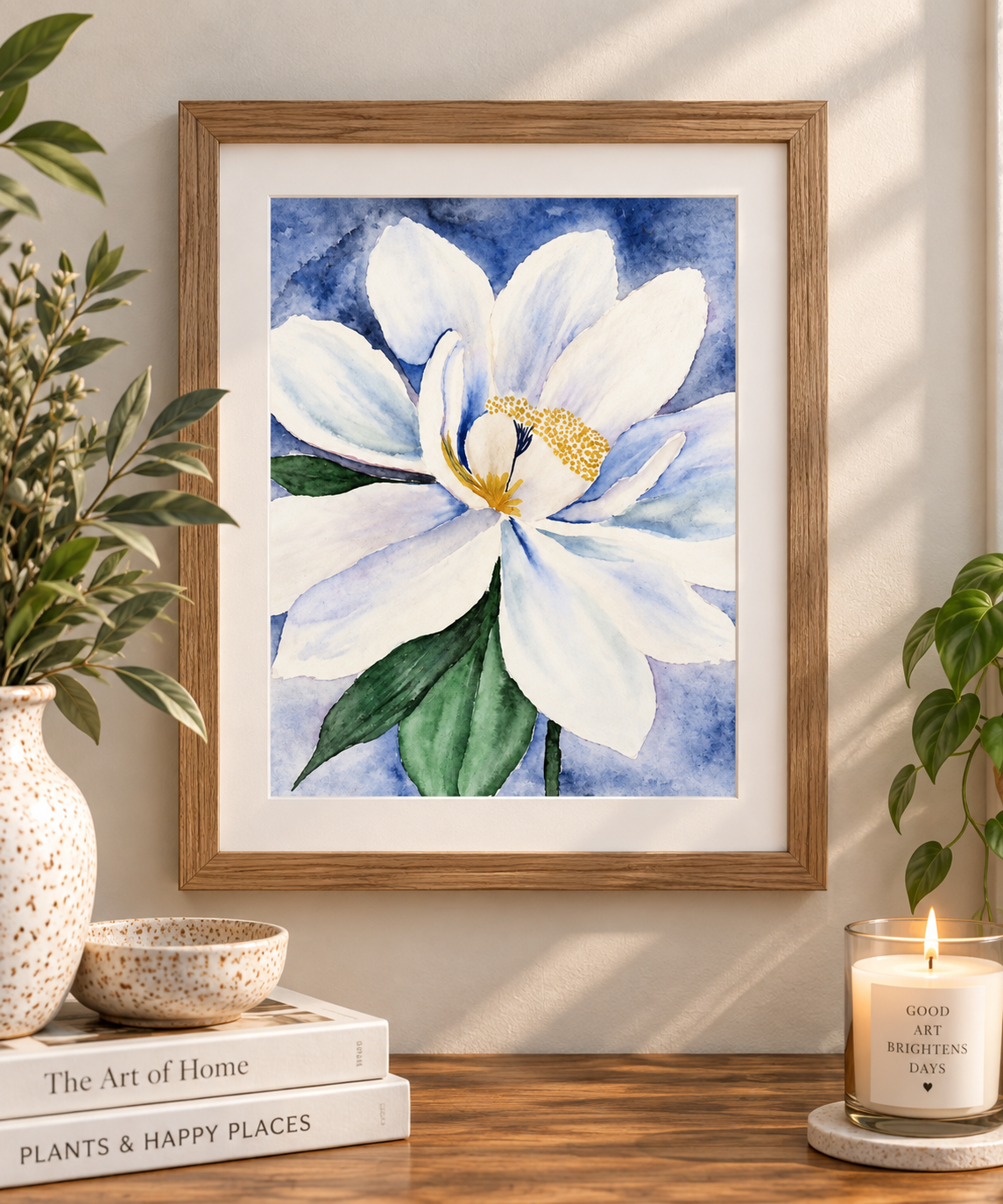 Framed watercolor print of a large white magnolia with soft blue and lavender shading, green leaves, and a textured blue background, displayed in a natural wood frame on a warm neutral wall.