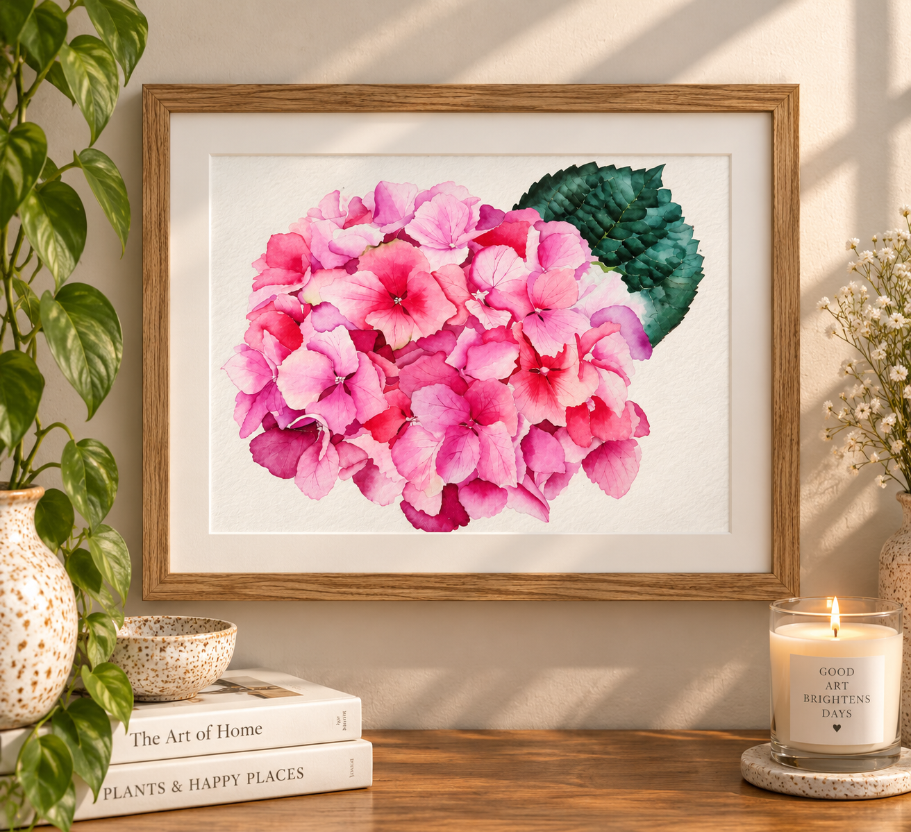 Framed watercolor print of vibrant pink hydrangea blooms with a deep green leaf, displayed in a natural wood frame on a warm neutral wall.