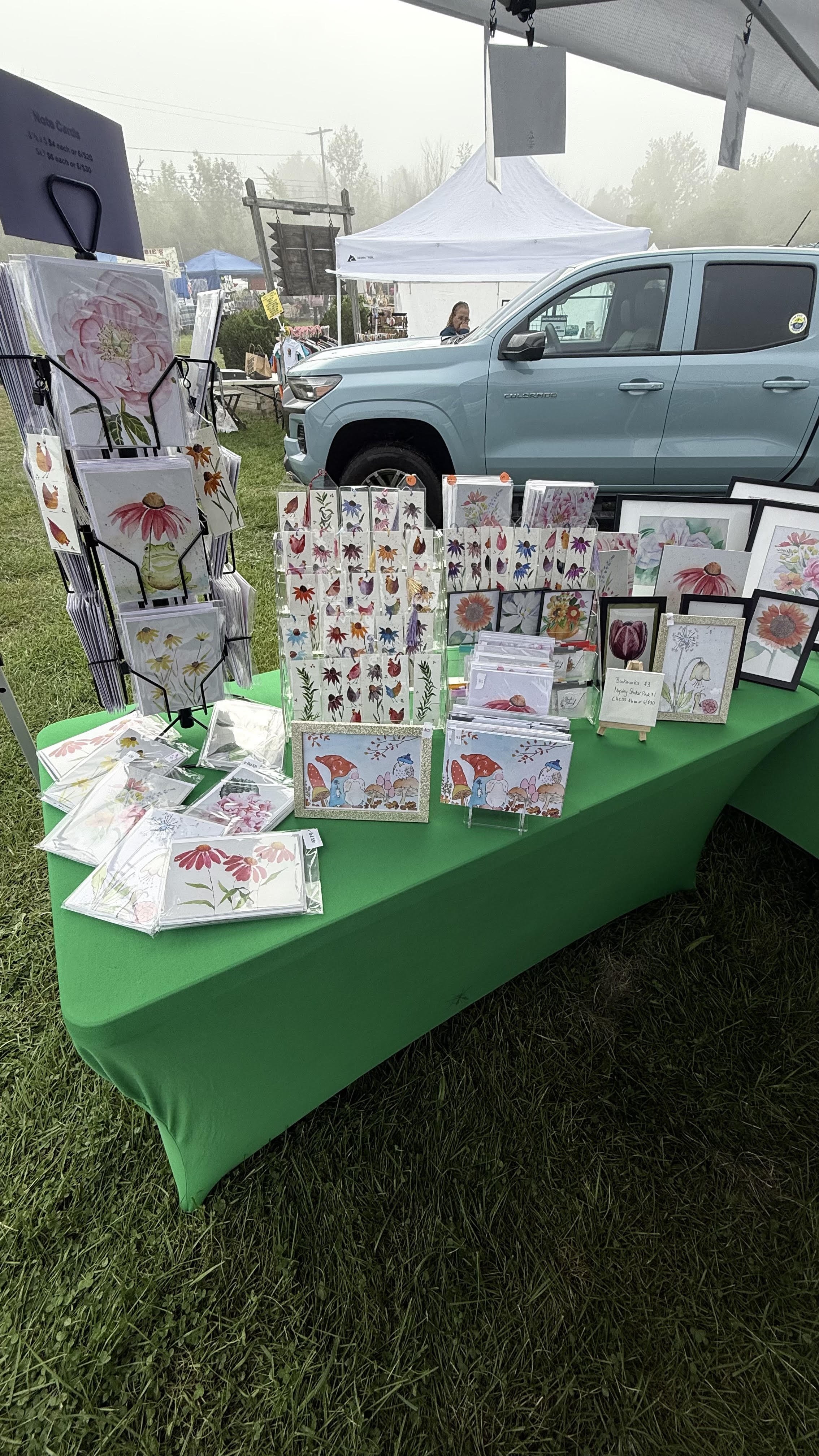 A display table with floral-themed greeting cards at an outdoor market, with a green tablecloth and a background of tents, a blue truck, and foggy weather.
