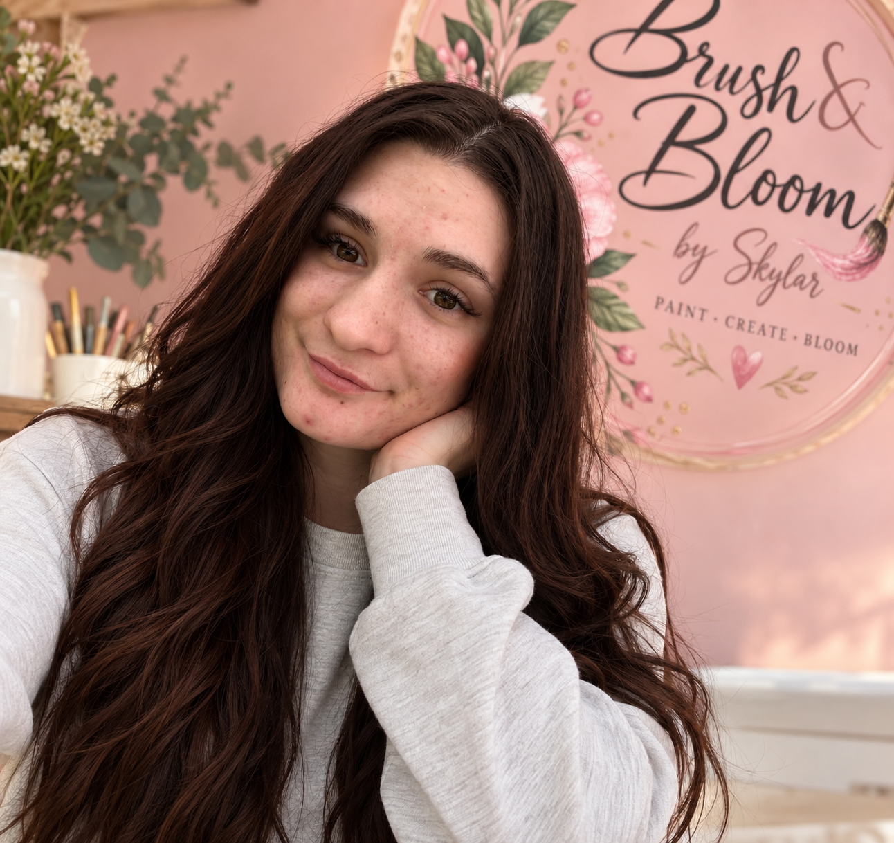 Young woman with long brown hair smiling in a pink art studio, with a Brush & Bloom by Skylar floral sign and art supplies in the background.