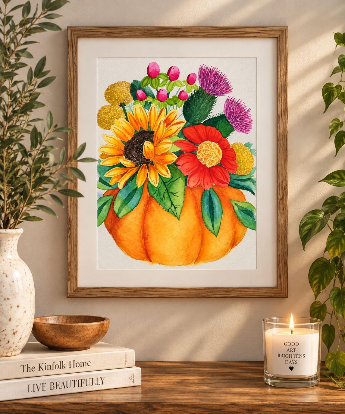 Framed watercolor print of a vibrant autumn bouquet with a sunflower, red and purple flowers, and greenery arranged in an orange pumpkin, displayed on a warm neutral wall.