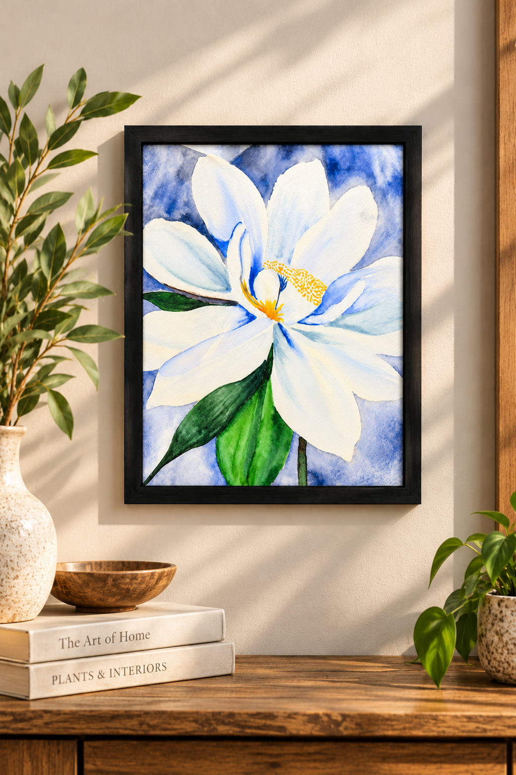 8x8 black framed watercolor print of a white magnolia with soft blue details, green leaves, and a blue watercolor background, displayed on a warm neutral wall.