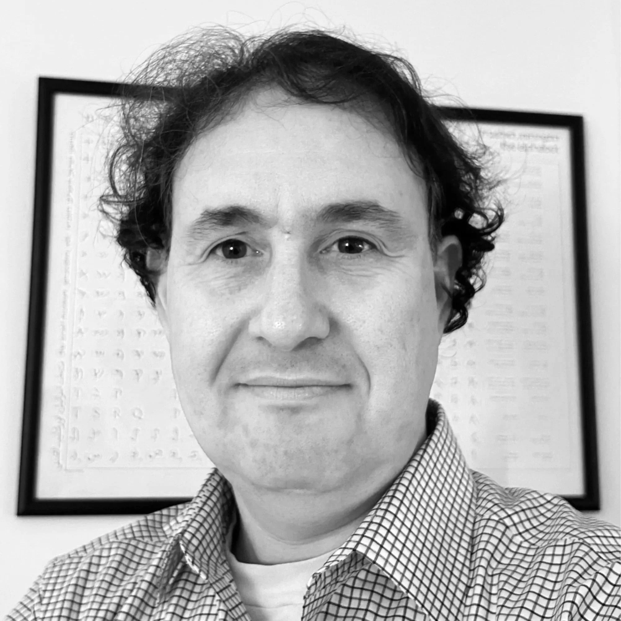 Black and white photo of a person with curly hair and wearing a checkered shirt, standing in front of a framed picture on the wall.