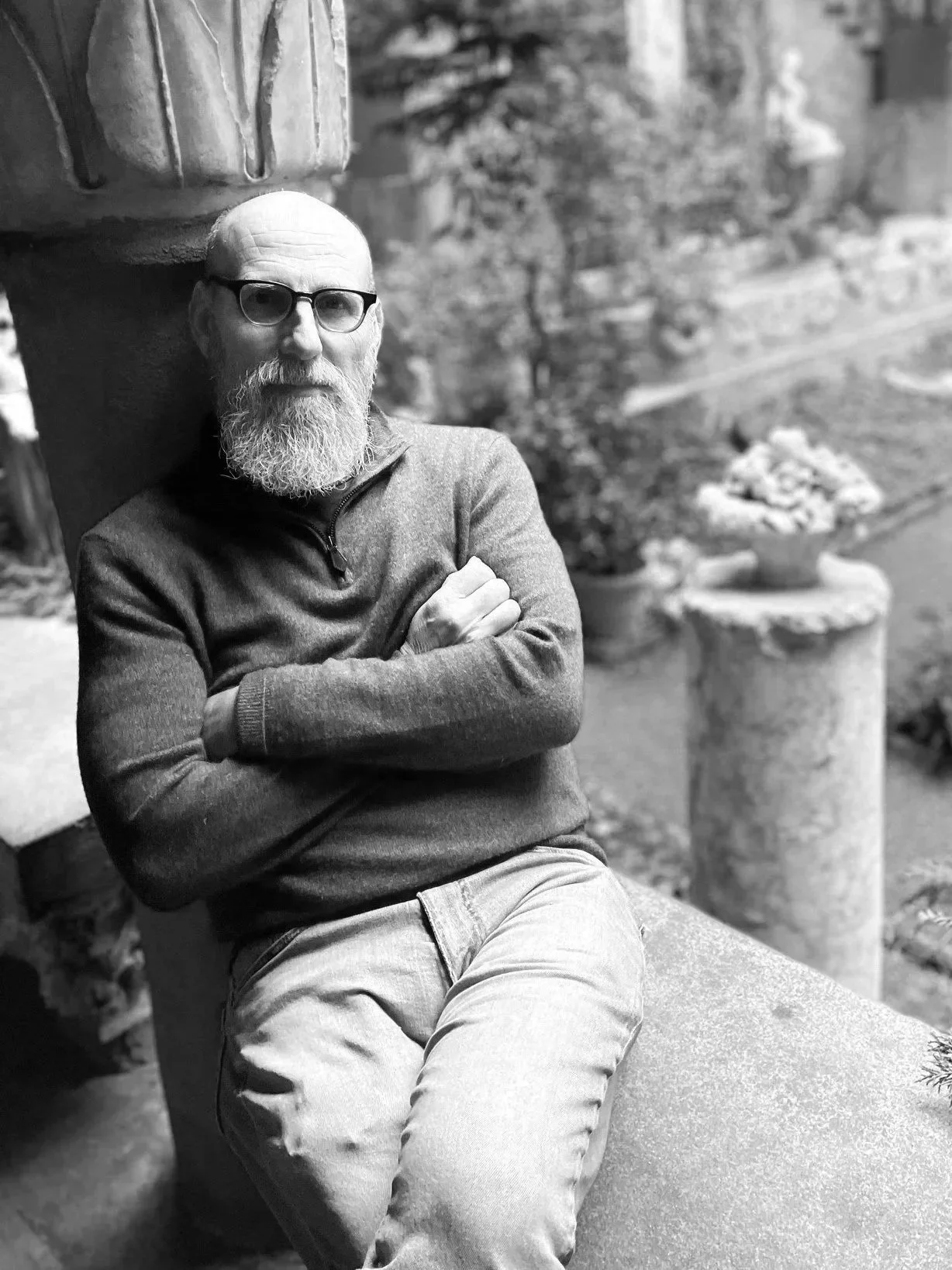 A black and white photo of a man with a white beard, glasses, wearing a sweater and light pants, sitting on a bench outdoors with crossed arms.
