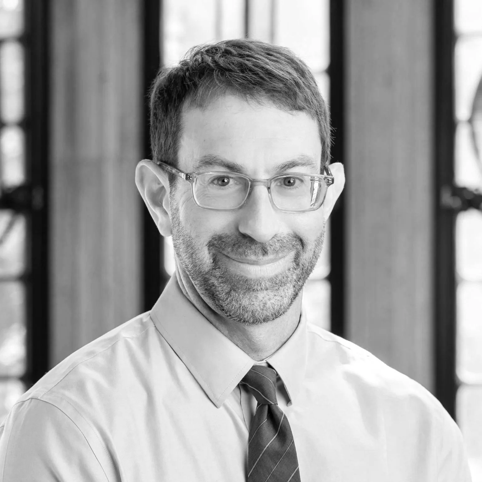 A black and white portrait of a smiling man wearing glasses, a shirt, and a tie, with a background of vertical and diagonal window panes.