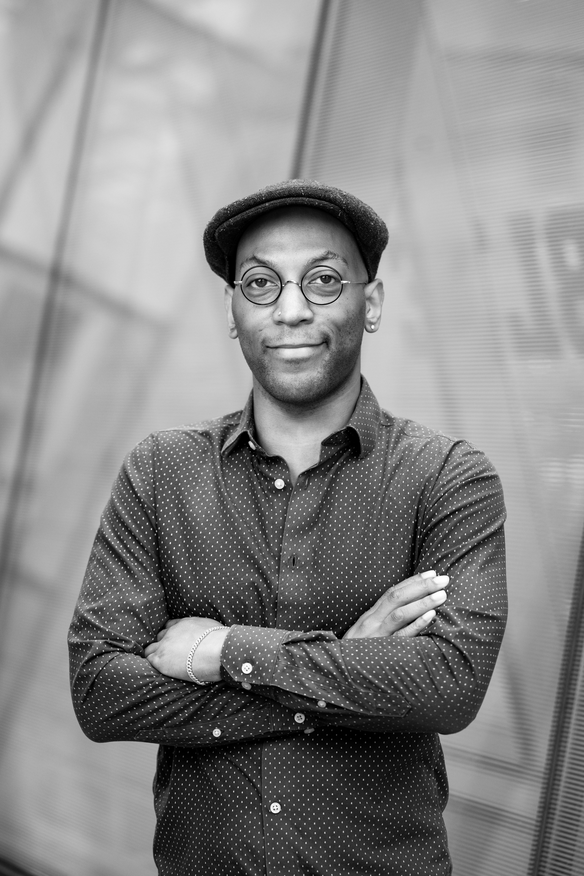 A man wearing glasses, a dark newsboy cap, and a polka dot shirt, standing with folded arms in front of a metallic wall or structure in a modern setting.