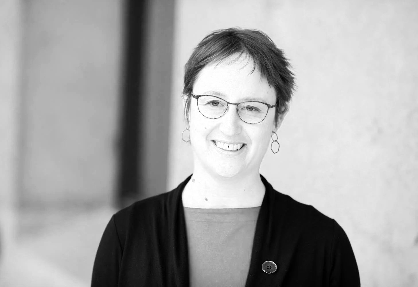 Black and white photo of a woman with short dark hair, glasses, and earrings, smiling, wearing a dark jacket over a light-colored top, with a blurred background.
