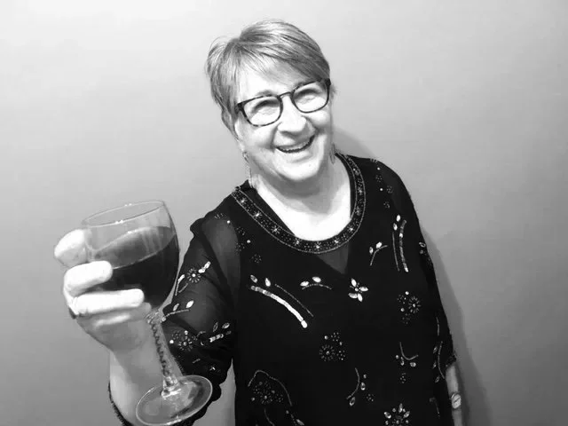A smiling woman with short hair and glasses holding a glass of red wine, dressed in a black top with decorative patterns, against a plain background.