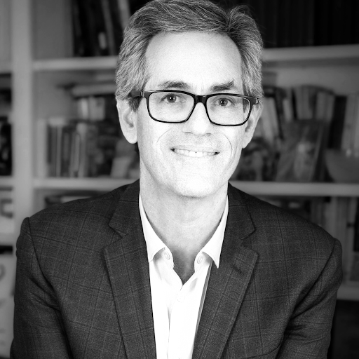 Black and white photo of a man with glasses, smiling, in front of bookshelf