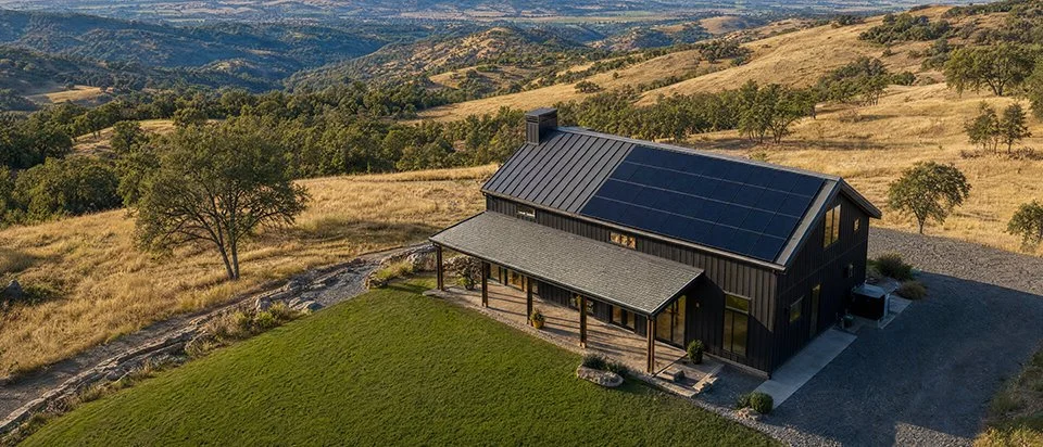 A black house with solar panels on the roof, surrounded by rolling hills and trees, with a green lawn in the front.