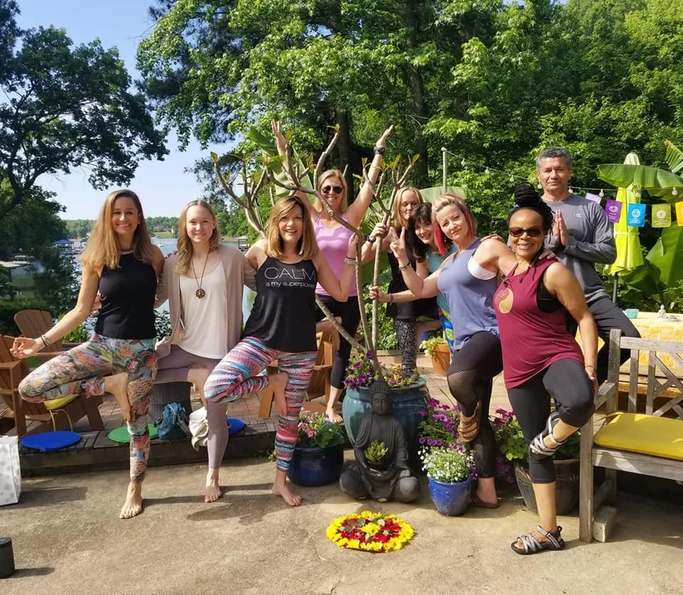 Group of nine people practicing yoga outdoors with greenery and a lake in the background, some performing tree pose, others in different yoga poses, surrounded by flowers and decorative items.