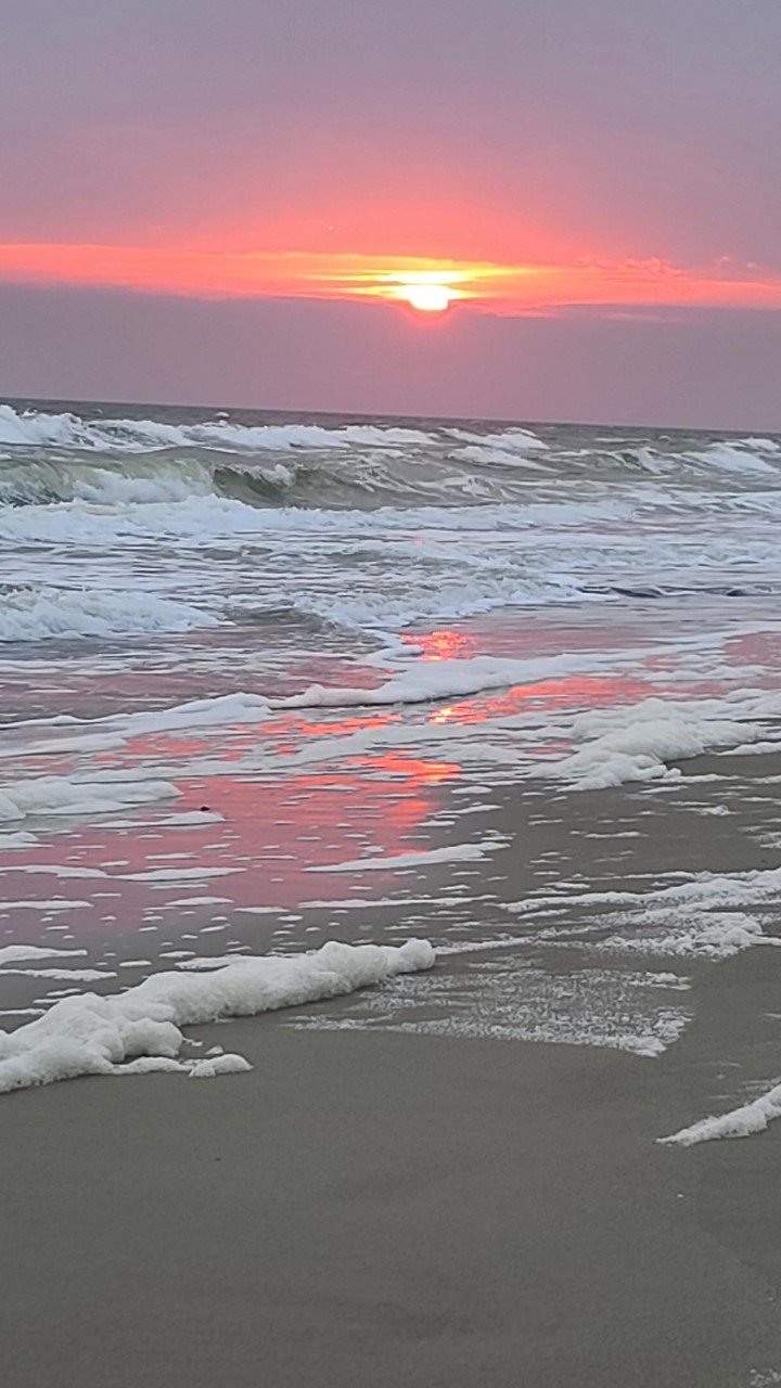 Sunset over the ocean with pink, orange, and purple sky and gentle waves washing onto the sandy beach, reflecting the colorful sky in Oak Island, NC