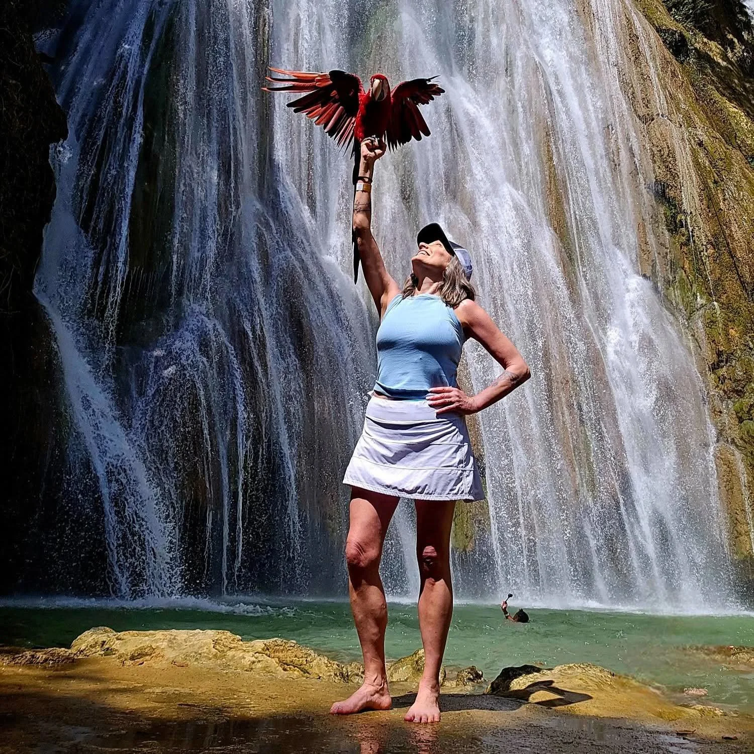 A woman in a light blue tank top, white skirt, and baseball cap stands on rocks in front of a waterfall, raising her left arm with a red exotic bird perched on her hand.